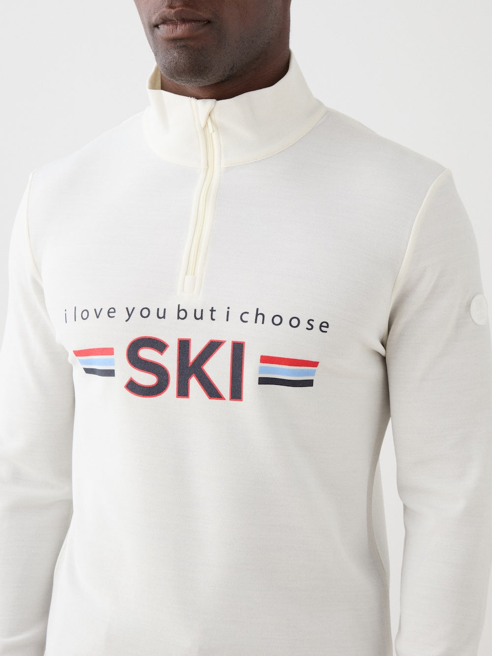 A person wears the We Norwegians Moore ZipUp men’s sweater, off-white with "i love you but i choose SKI" and red, white, and blue retro ski base layer-inspired stripes on the front.