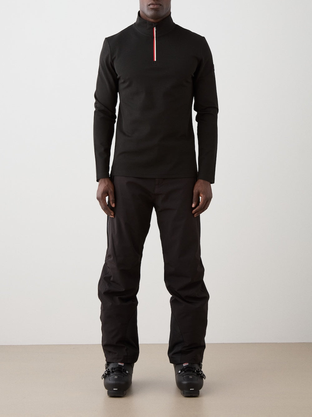 A person stands facing forward against a plain background, wearing the We Norwegians Moore ZipUp Solid men’s black base layer with a partially red zip, paired with black pants and black ski boots.