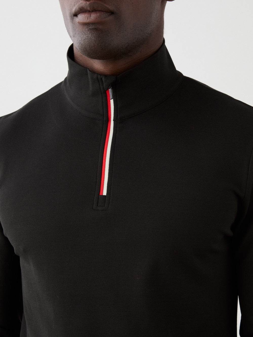 A man wears the We Norwegians Moore ZipUp Solid—a black alpine-style base layer with a high collar, half-zip, and red, white, and black striped trim; only his lower face and upper torso are visible.