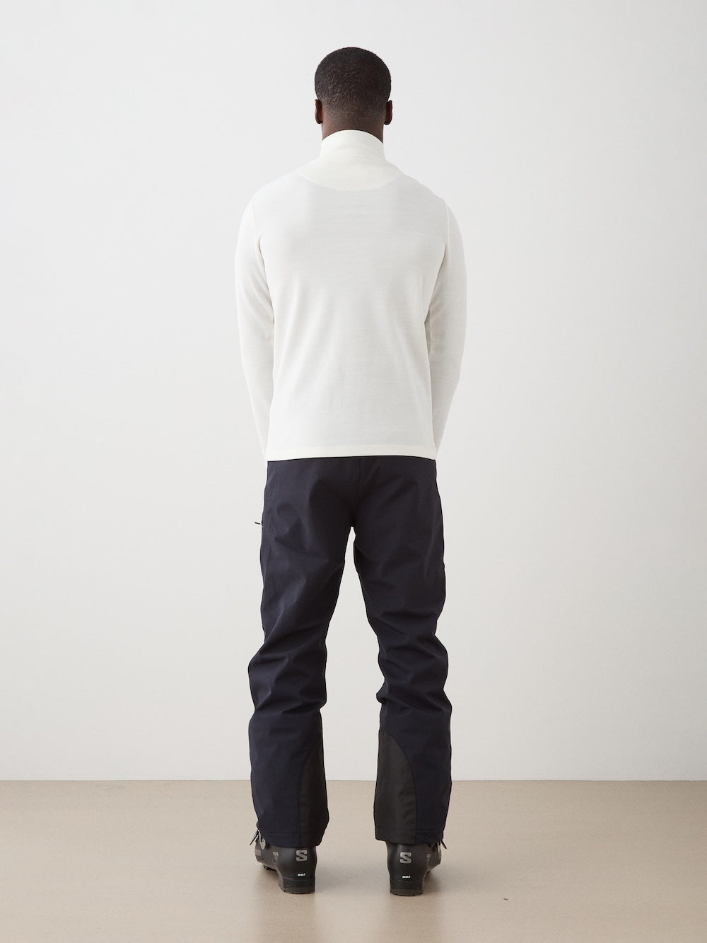 A person stands facing away from the camera wearing the We Norwegians Moore ZipUp Solid men's white Merino wool long-sleeve top, dark pants, and black boots against a plain light-colored background.