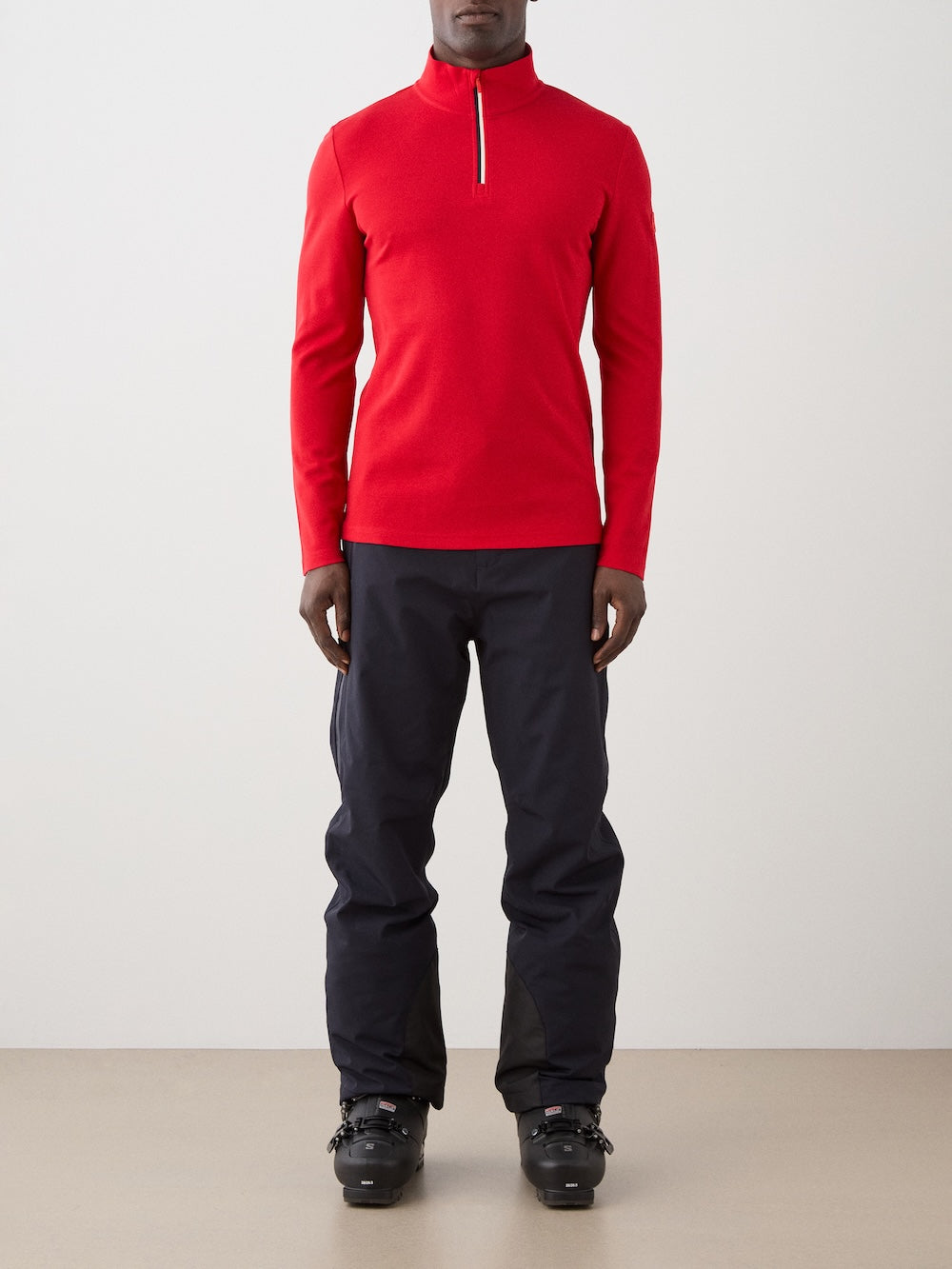 A man faces forward against a plain light background, wearing the We Norwegians Moore ZipUp Solid men in bright red, paired with black pants and black ski boots.