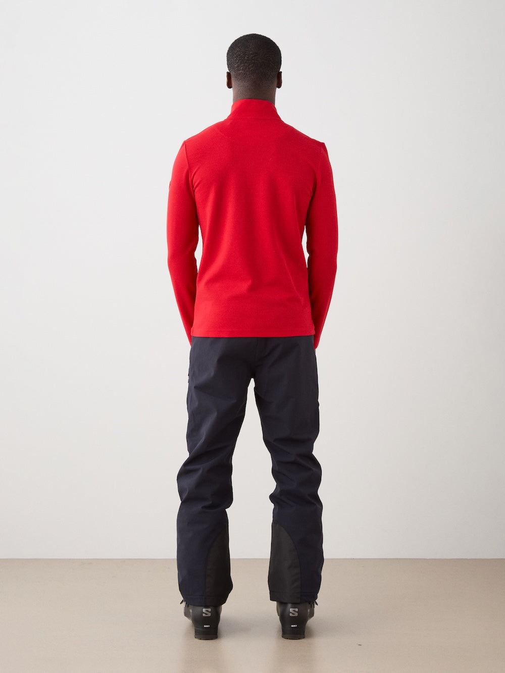 A person faces away from the camera, wearing a We Norwegians Moore ZipUp Solid men’s long-sleeve red top, dark pants, and black shoes, set against a plain light background.