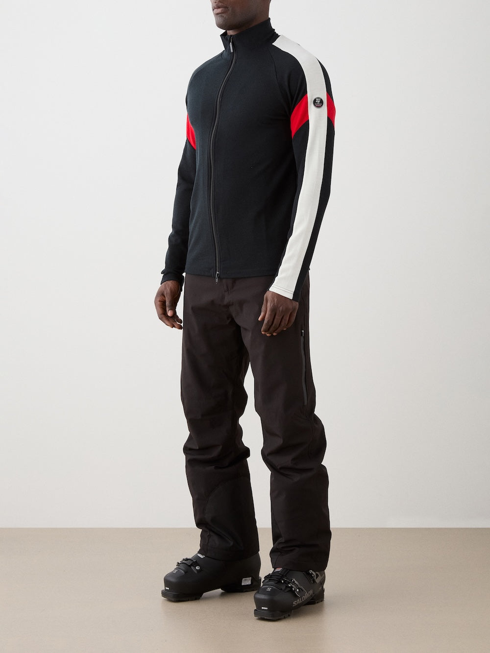 A man stands against a plain background wearing the We Norwegians Zermatt FullZip men black jacket with white and red sleeve stripes, paired with black pants and ski boots. Ideal as a ski mid-layer, this Merino wool jacket delivers warmth and style.