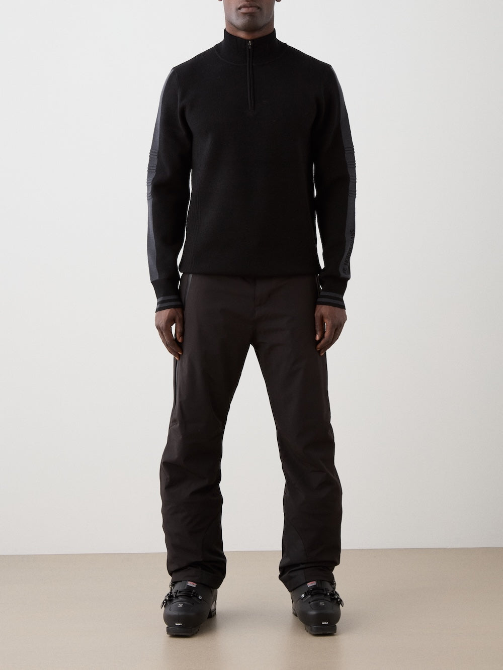 A man stands facing forward in a We Norwegians Rocky ZipUp men sweater, crafted from black merino wool, paired with black pants and ski boots against a simple light background, arms relaxed at his sides.