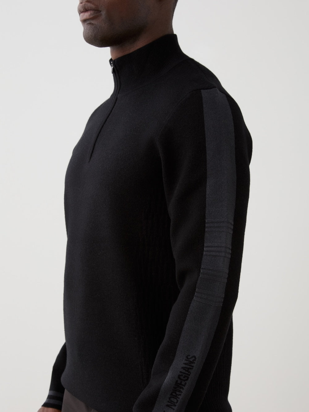 A man wears the We Norwegians Rocky ZipUp men, a black long-sleeve sweater with a dark stripe and subtle sleeve lettering, inspired by vintage alpine style, standing against a plain light background.
