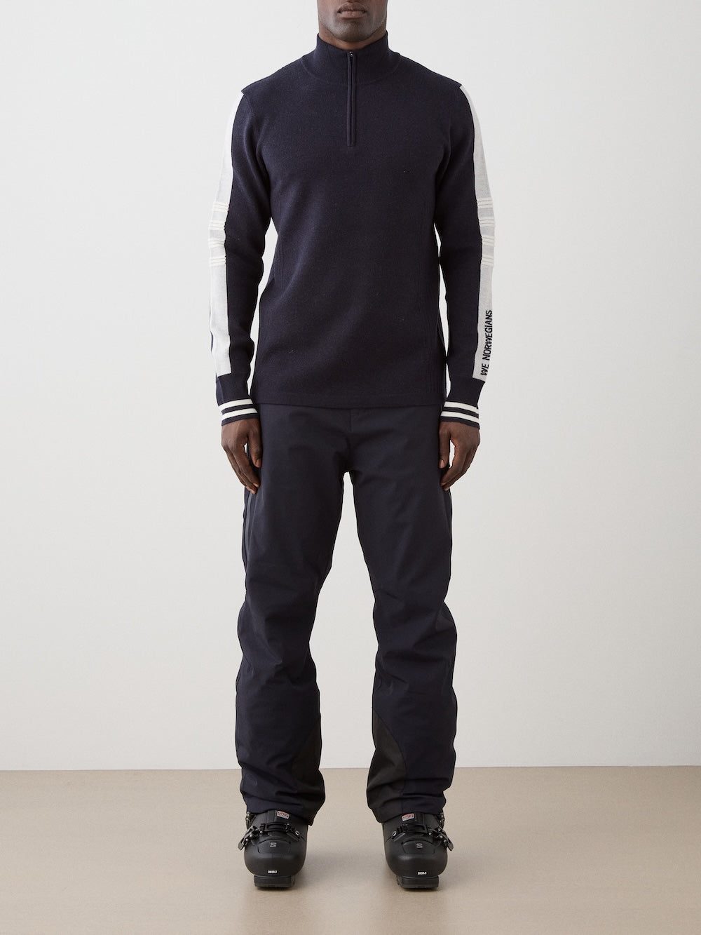 A person stands facing forward in a dark We Norwegians Rocky ZipUp men with white sleeve stripes, black pants, and black ski boots, channeling vintage alpine style against a plain background.