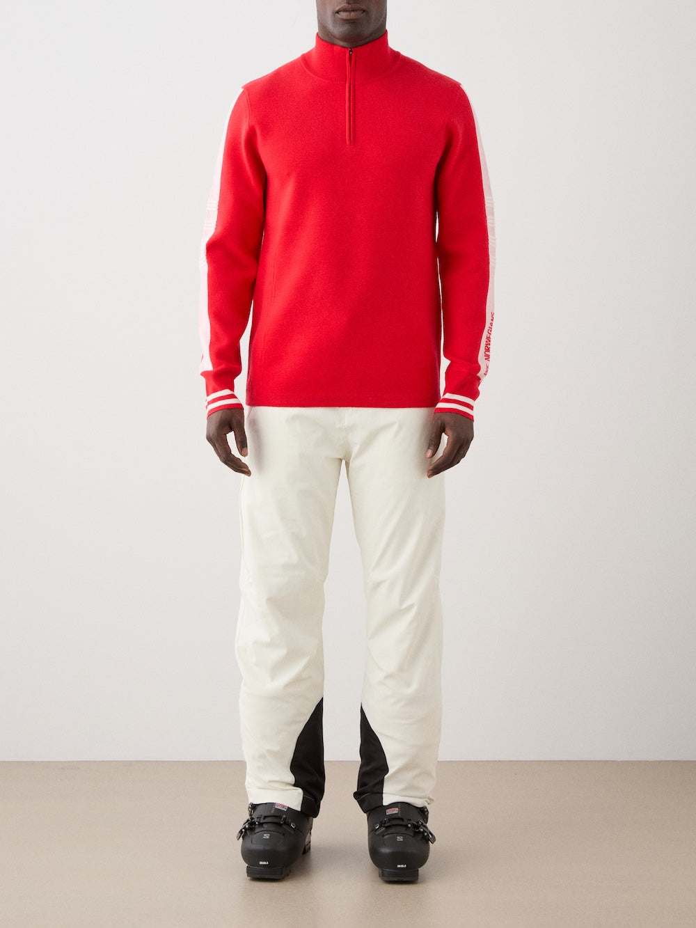 A person faces forward in the We Norwegians Rocky ZipUp men sweater with white sleeve stripes, evoking vintage alpine style. They're paired with white pants featuring black ankle patches and black ski boots, against a plain light backdrop.