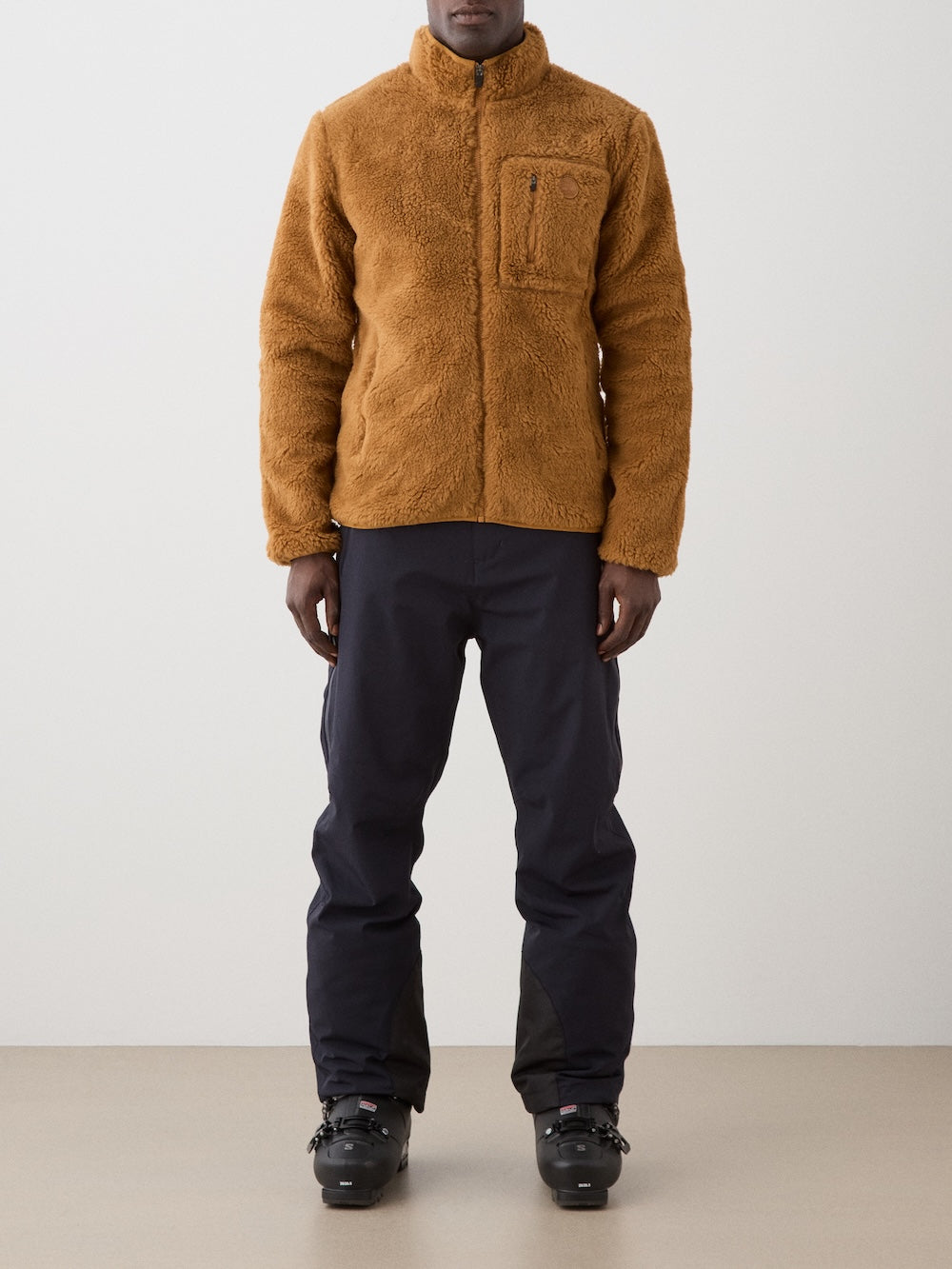 A person wearing a We Norwegians Kebnekaise Jacket men in brown, with a zippered chest pocket and high collar, along with dark pants and black ski boots, stands indoors against a plain background. The person's face is not visible.