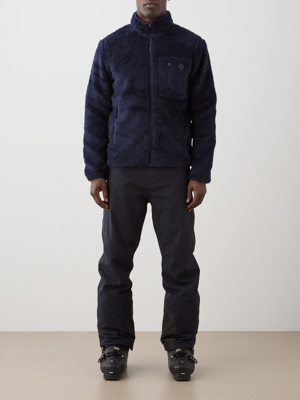 A person, face not visible, wears a We Norwegians Kebnekaise Jacket men in navy blue, paired with black pants and ski boots against a plain background, suitable for cold weather or winter sports.