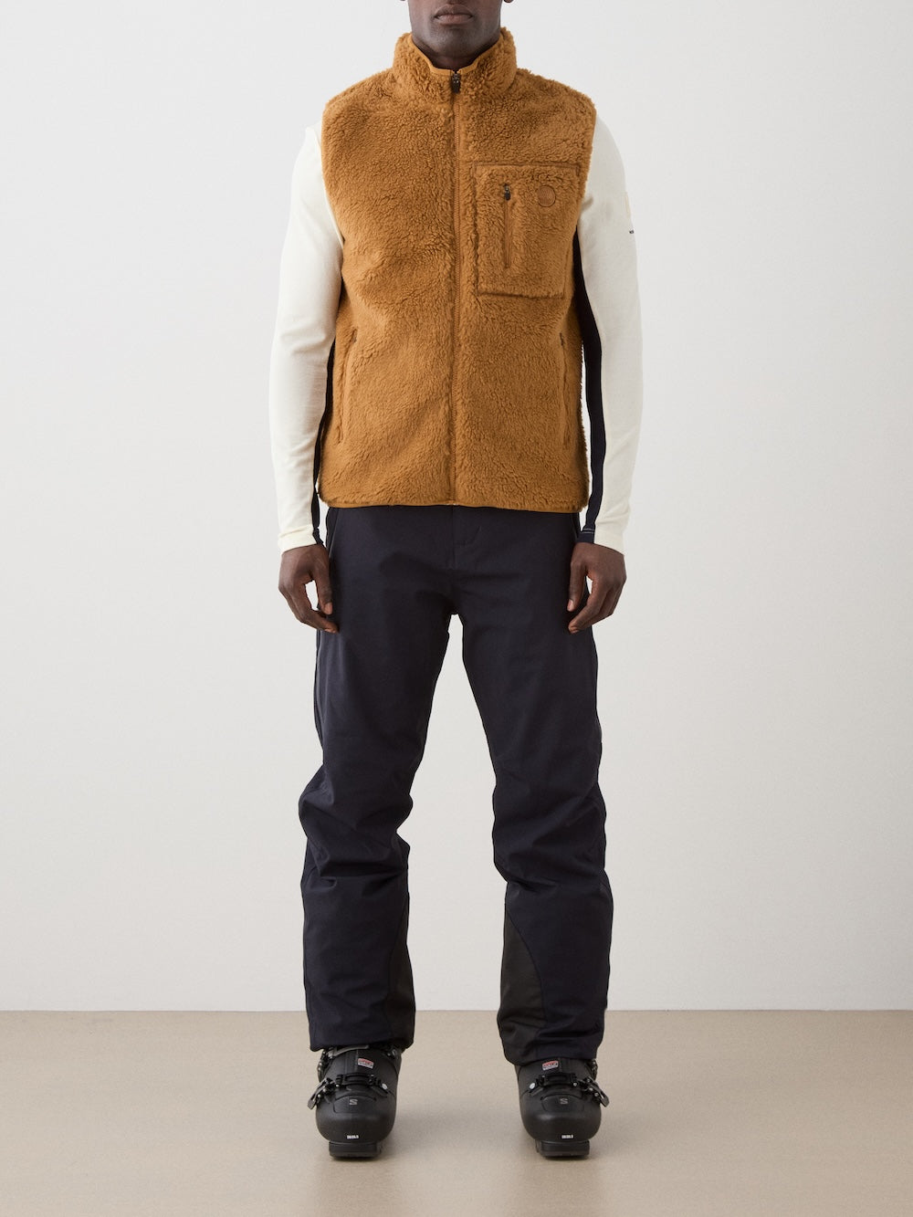 A man stands indoors, facing the camera against a plain light background, wearing the We Norwegians Kebnekaise Vest men—a tan wool-blend pile vest—over a long-sleeve white top with dark pants and black ski boots.