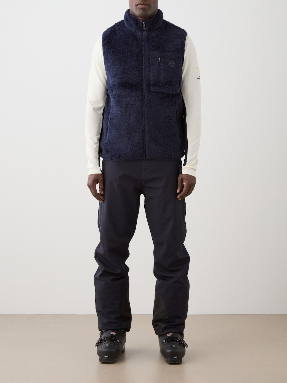 A man faces forward wearing the We Norwegians Kebnekaise Vest men—a navy wool-blend pile vest perfect as an après-ski mid-layer—styled over a cream long-sleeve top, black pants, and black ski boots against a light background.