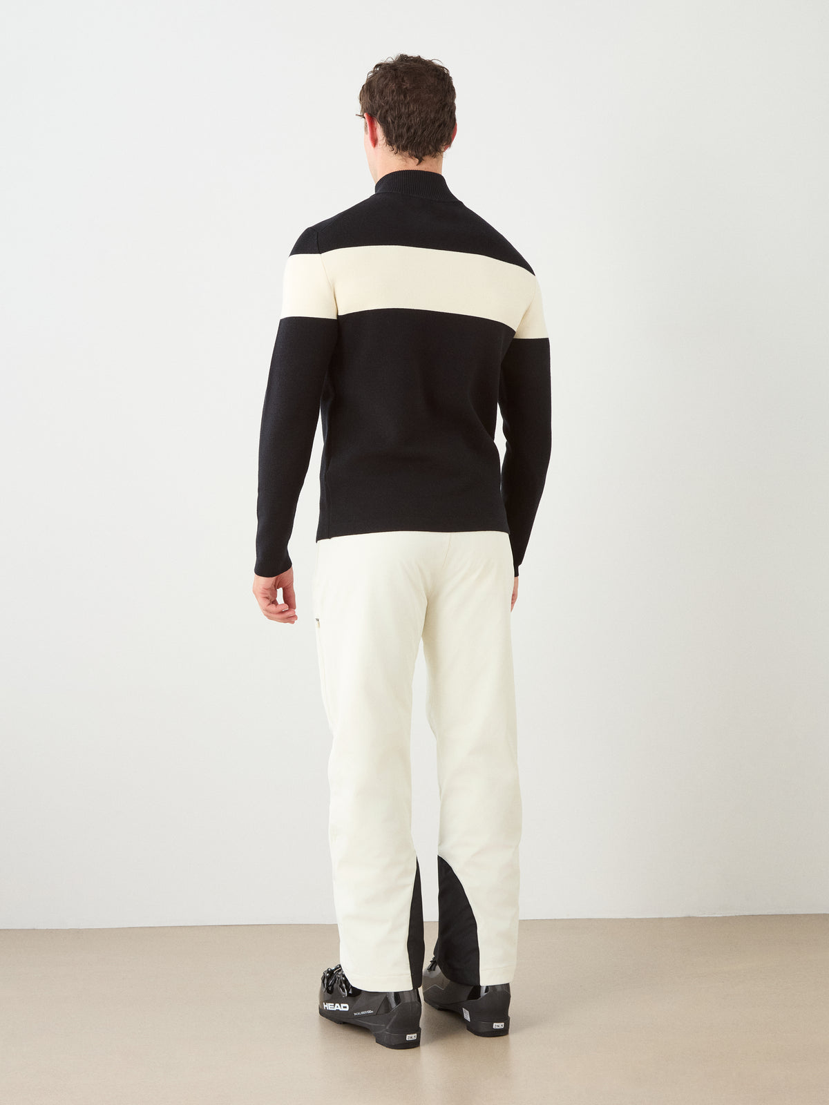 A person stands facing away, wearing the We Norwegians Holmenkollen ZipUp men sweater in black and white, paired with white pants and black ski boots, against a plain light background.