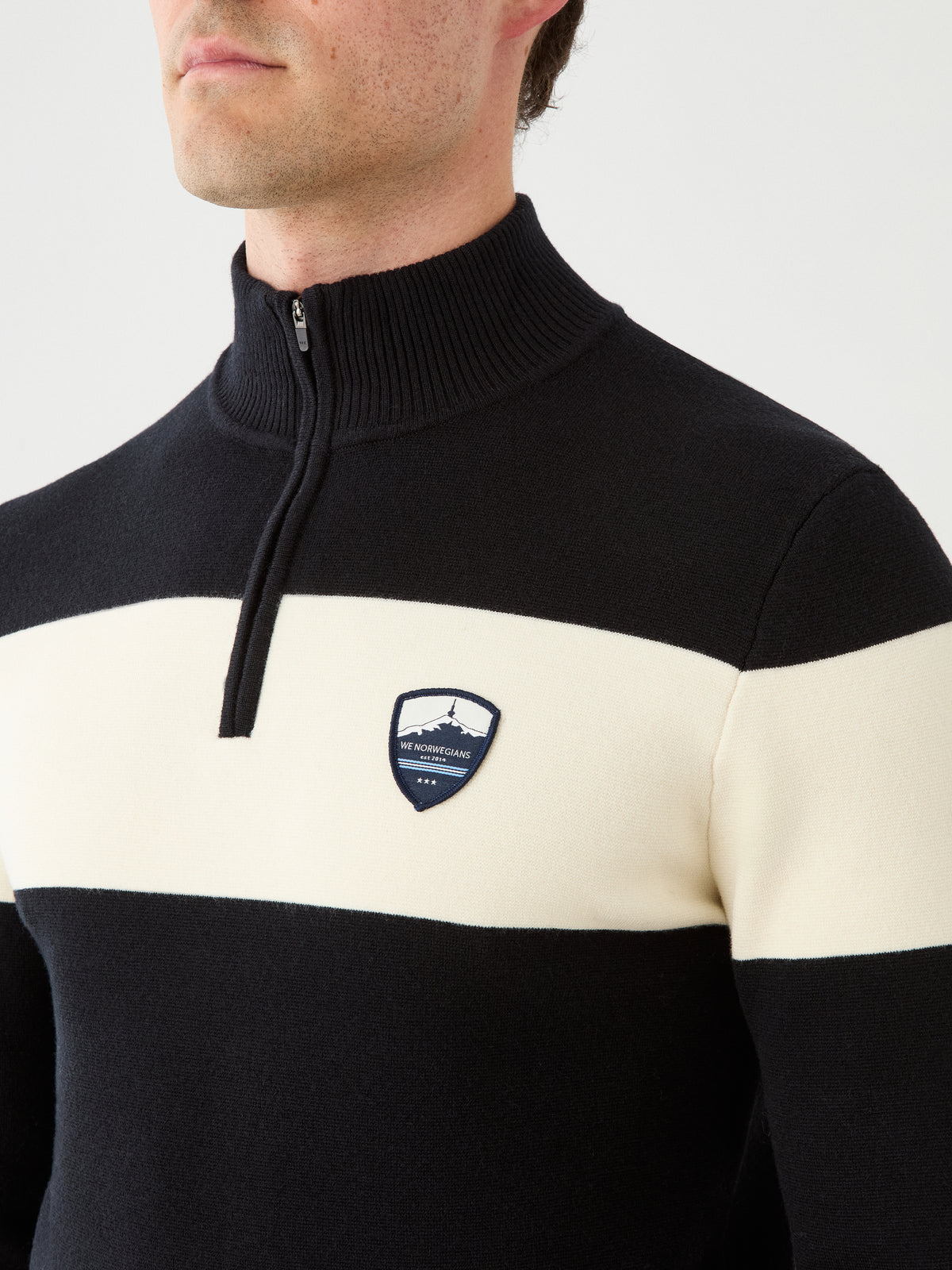 A man wearing the We Norwegians Holmenkollen ZipUp men sweater in a black and white striped pattern, featuring a high collar and a crest logo on the chest reminiscent of a vintage ski sweater, shown from shoulders to mid-face against a plain background.