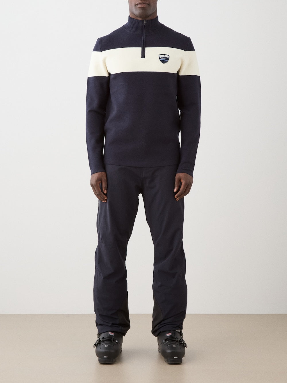 A person stands indoors against a plain, light background wearing the We Norwegians Holmenkollen ZipUp men sweater in navy and white, featuring a front zipper and chest logo, paired with black pants and ski boots.
