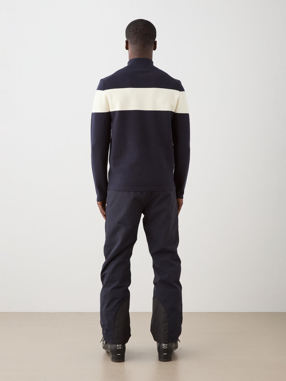 A man faces away from the camera, wearing the We Norwegians Holmenkollen ZipUp men—a navy merino wool sweater with a white stripe across the upper back—and matching navy pants, indoors on a beige floor against a plain light wall.