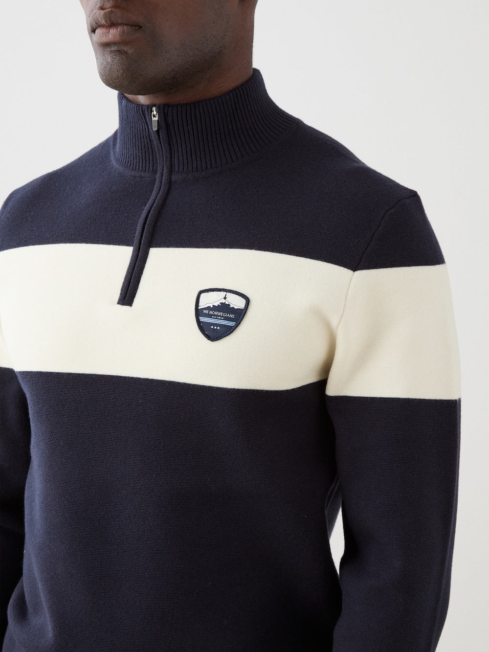 A person wears the We Norwegians Holmenkollen ZipUp men sweater in navy blue and white with a horizontal white stripe and crest patch on the chest. The face is partially out of frame.