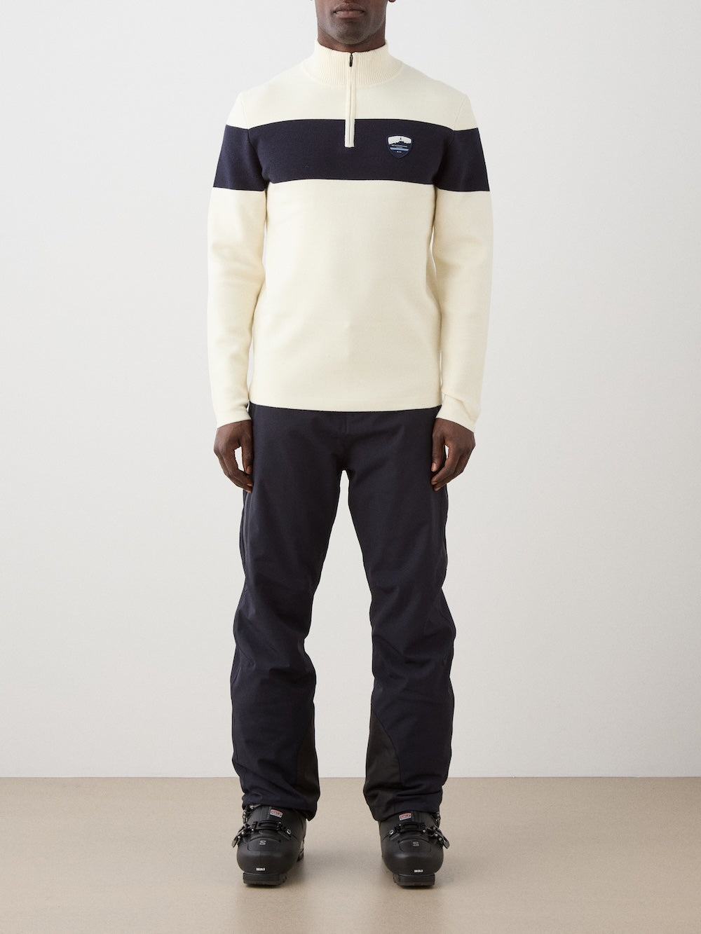 A person wears the We Norwegians Holmenkollen ZipUp men sweater in cream and navy with a small chest logo, paired with navy pants and black ski boots, standing against a plain light background.