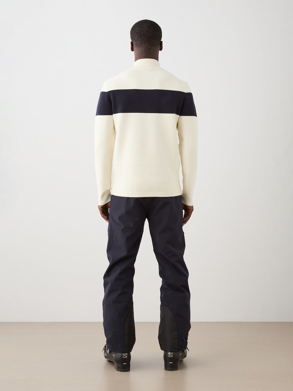 A man faces away from the camera, wearing the We Norwegians Holmenkollen ZipUp men—a cream merino wool sweater with a wide black stripe across the upper back—and black pants, set against a plain light background.