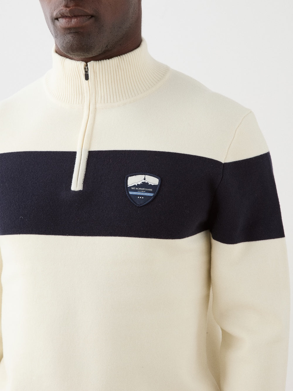 A man wears the We Norwegians Holmenkollen ZipUp—a cream sweater with a black chest stripe, zip collar, and left-side patch—showcasing a classic vintage ski style.