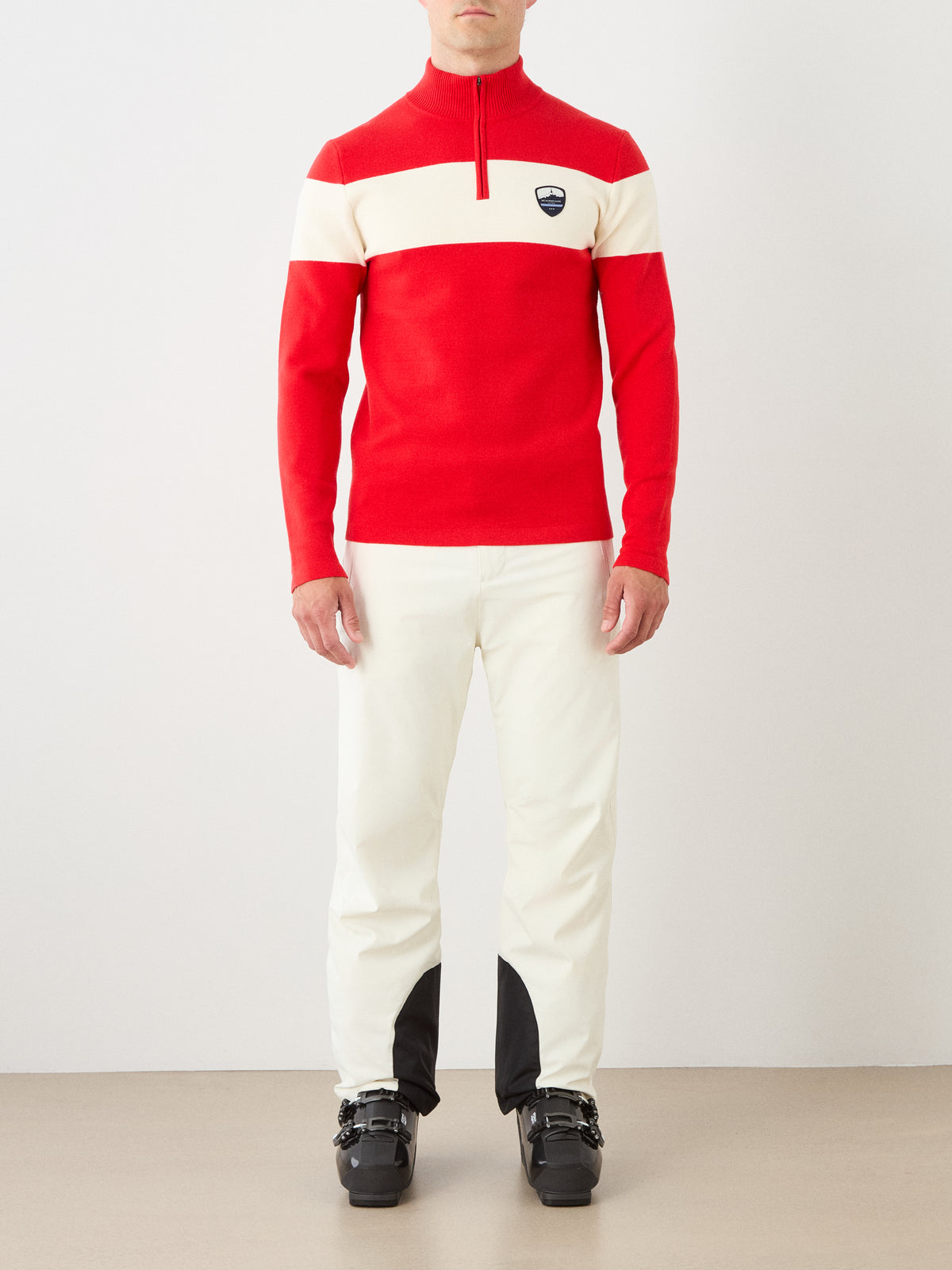 A person stands against a plain light background wearing the We Norwegians Holmenkollen ZipUp for men—a red and white vintage-inspired ski sweater—paired with white pants featuring black patches on the ankles and black ski boots.