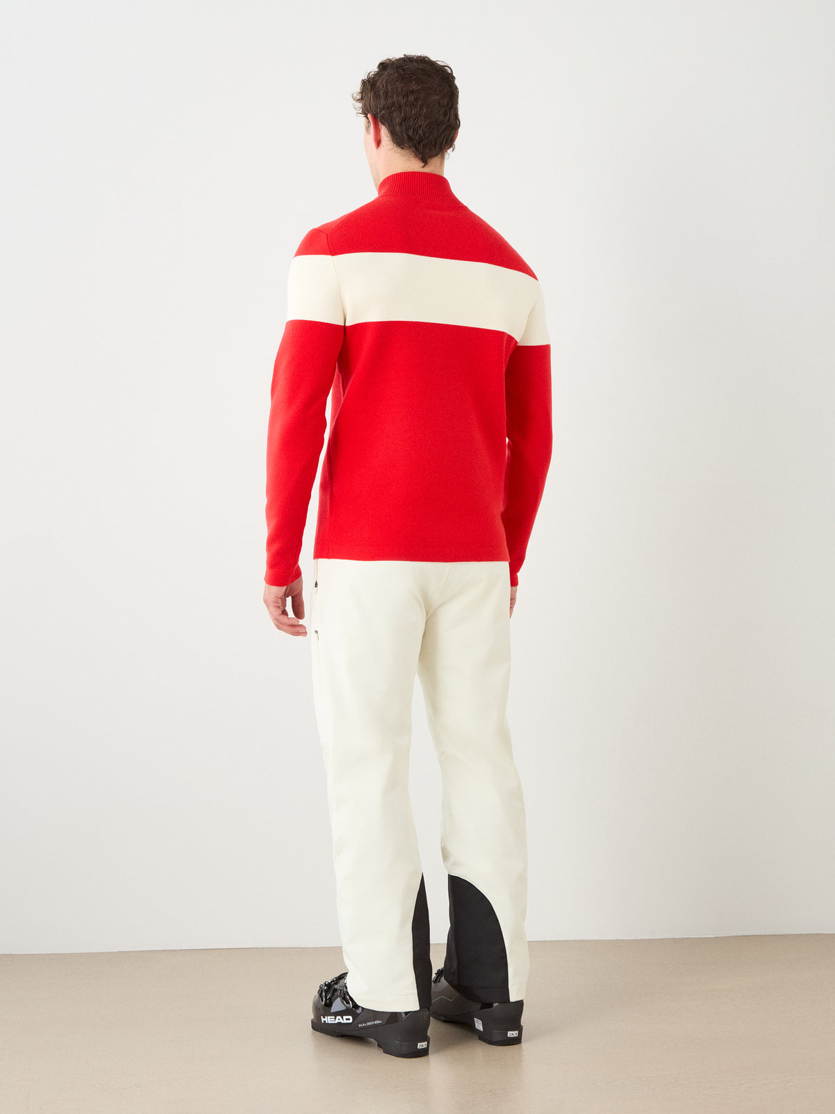 A person stands facing away in a minimal indoor setting, wearing the We Norwegians Holmenkollen ZipUp men—a vintage-style red ski sweater with a wide white horizontal stripe—paired with white pants featuring black patches near the ankles.