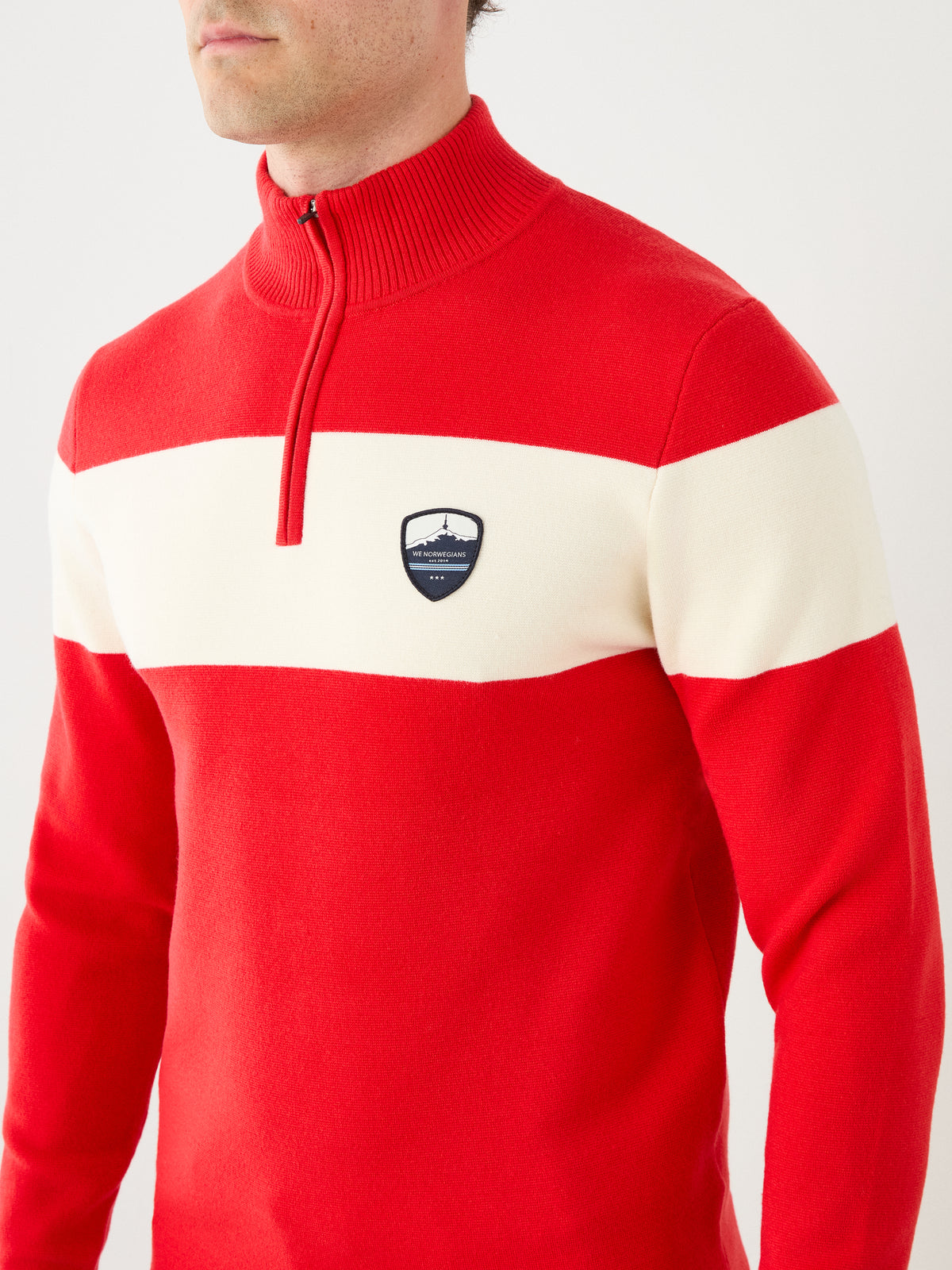 A person wearing the We Norwegians Holmenkollen ZipUp men sweater in red, featuring a white horizontal stripe, half-zip collar, and a small shield-shaped emblem on the chest. The person’s face is partially out of frame.