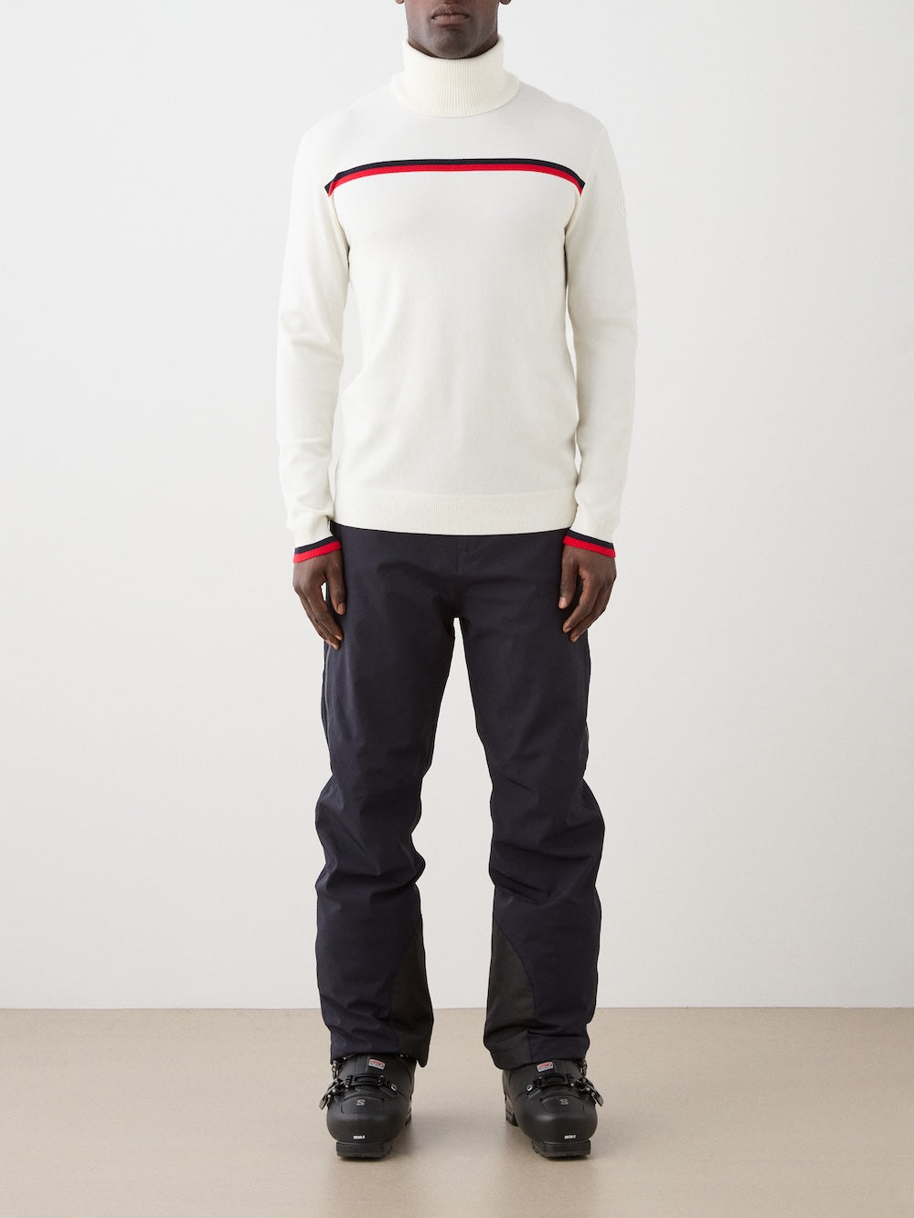 Person wearing a white sweater with red accents and dark pants on a plain background