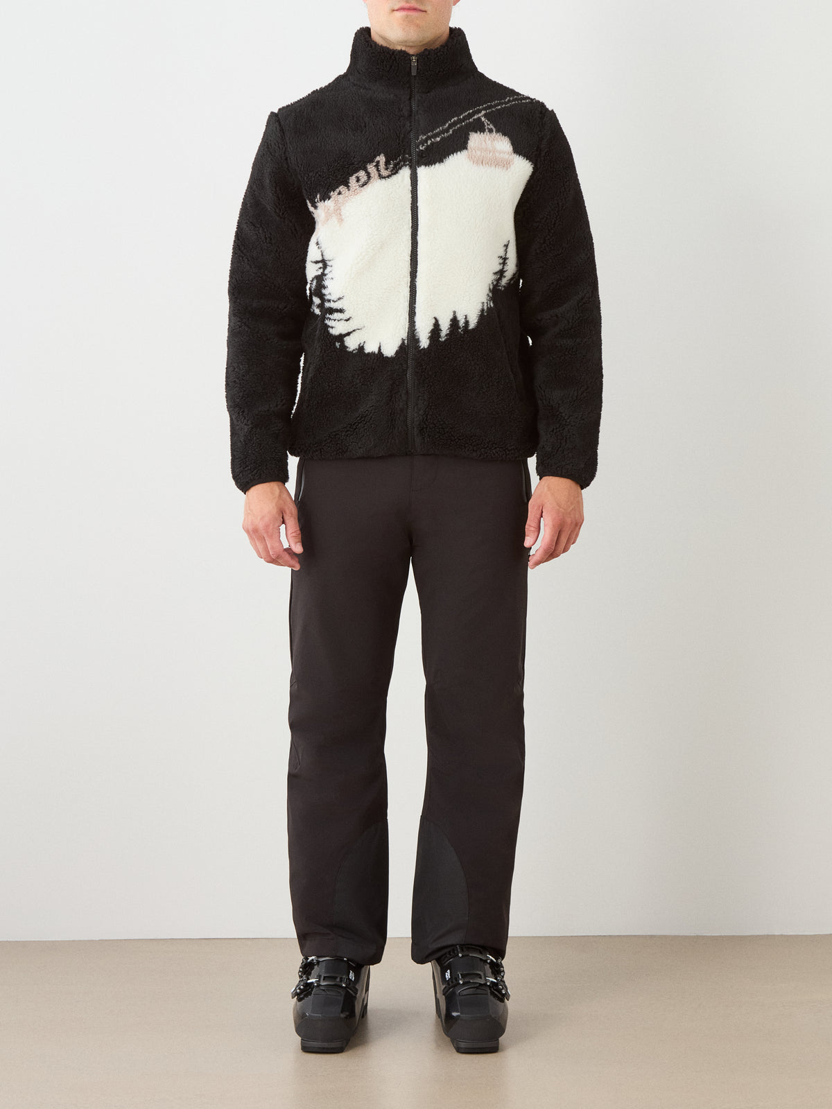 A man models the We Norwegians Snowmass Jacket—a black wool-blend piece showcasing a white ski lift scene—paired with black pants and boots, standing against a neutral background for an effortlessly stylish après ski look.