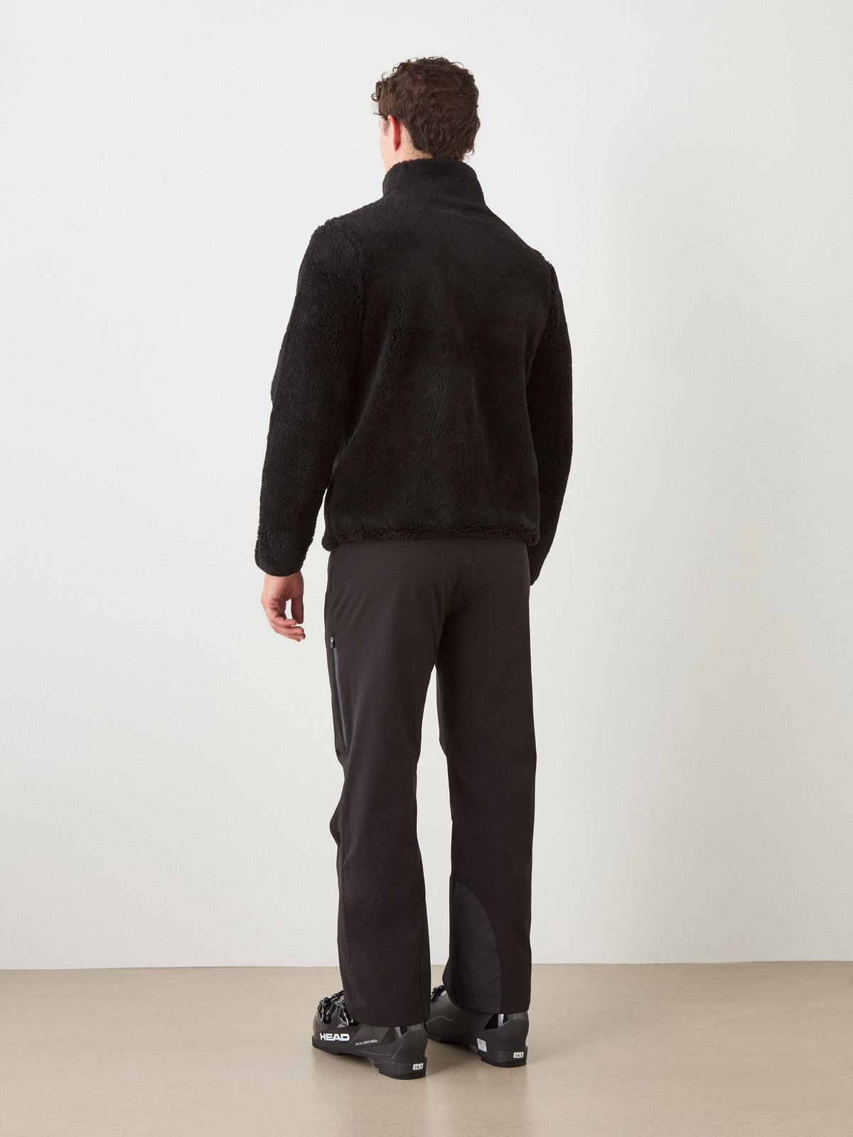 A person stands facing away, dressed in the We Norwegians Snowmass Jacket men—an elegant black wool-blend jacket perfect for après ski—paired with black pants and black boots featuring visible branding, set against a plain white wall and beige floor.