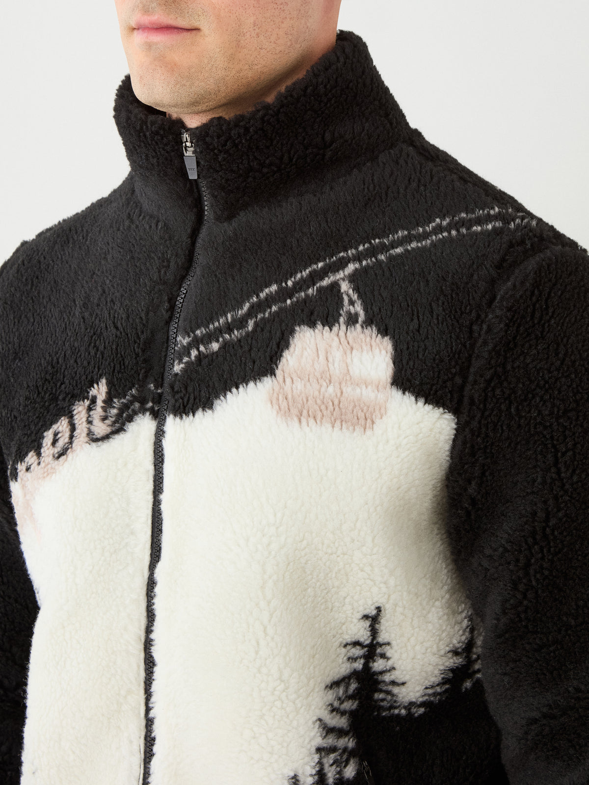 A person is seen wearing the We Norwegians Snowmass Jacket men, a zip-up black and white jacket featuring a high collar and an abstract mountain or ski lift design on the front. This stylish après ski piece from We Norwegians covers only the lower face and torso in the image.