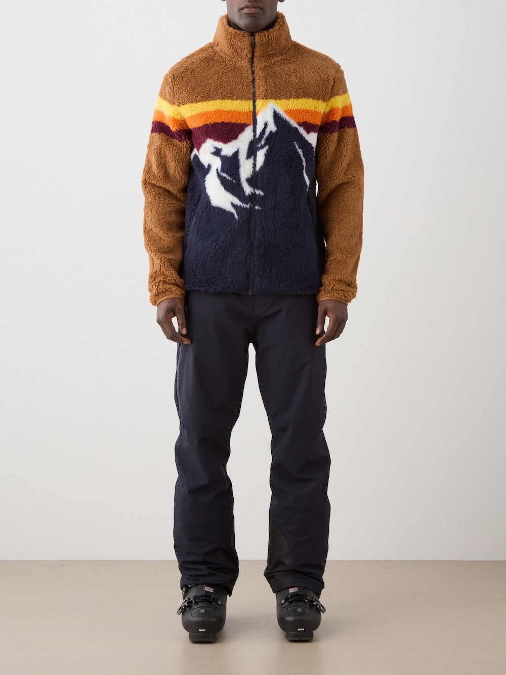 A person stands facing forward, wearing a We Norwegians Snowmass Jacket men—a brown and navy fleece with a mountain design and horizontal stripes—paired with dark pants and black winter boots, against a plain light background.