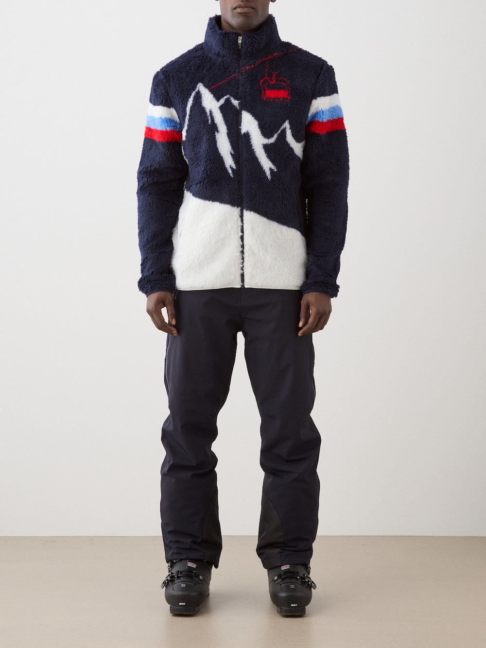 A person indoors wears We Norwegians' Snowmass Jacket men—an après ski piece in navy and white with mountain designs, red and blue sleeve stripes, a red ski lift graphic on the chest—paired with black ski pants and boots.