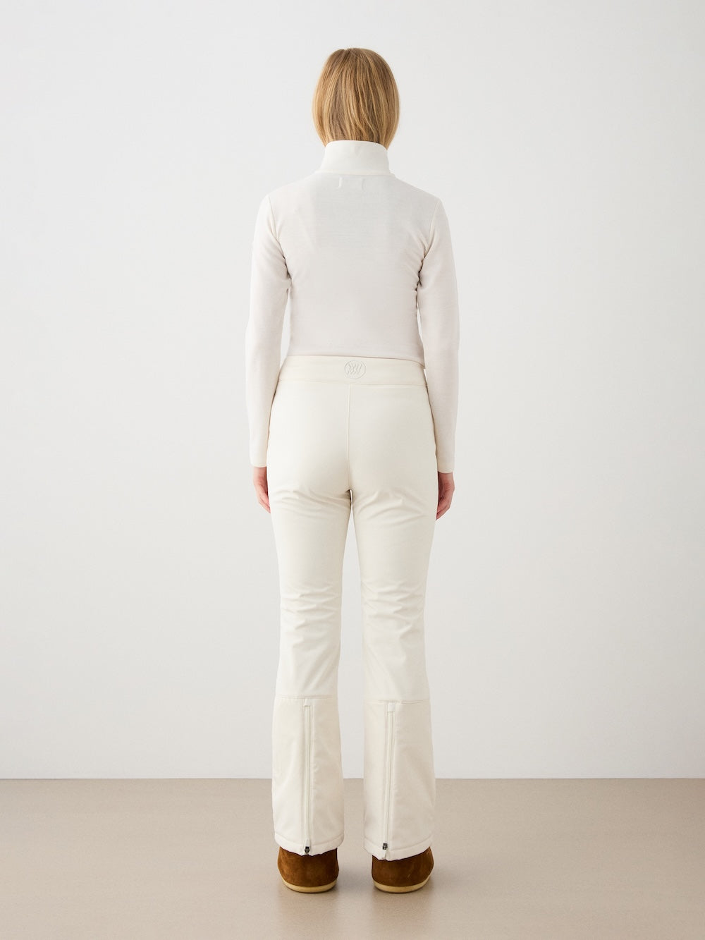 A woman with straight blonde hair faces away, wearing the We Norwegians Bibi ZipUp Solid women base layer in off-white and brown boots—ideal for a ski trip—against a simple light background.