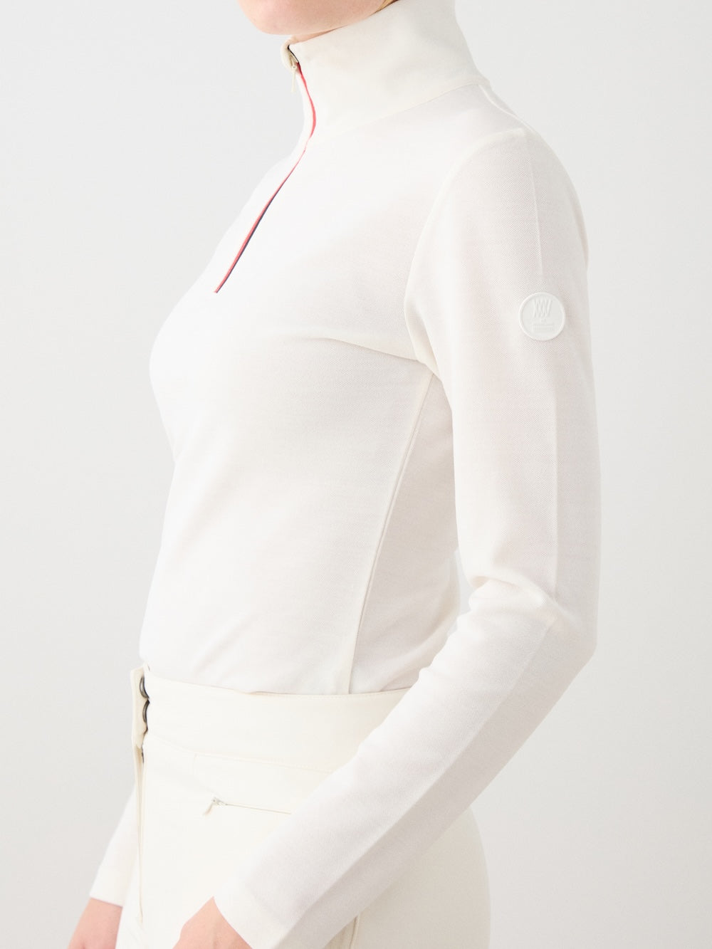 A woman wears the We Norwegians Bibi ZipUp Solid—a fitted, long-sleeve white Merino wool turtleneck with a red zipper—paired with white pants. Face partially out of frame; ideal as a stylish ski trip base layer.