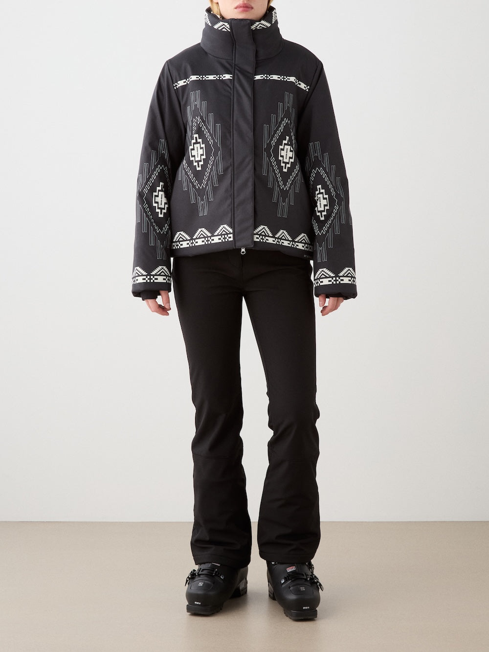 A person stands against a plain background wearing the black We Norwegians Pioneer Ski Jacket for women with white geometric patterns, paired with black ski pants and ski boots. The person’s face is partially out of frame.
