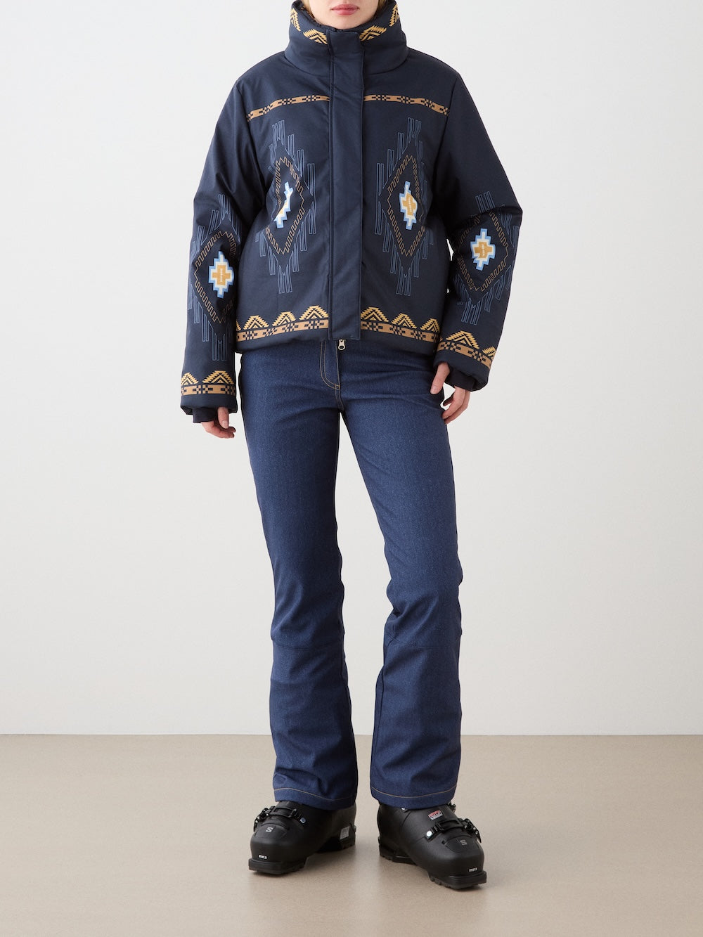 A person wears the We Norwegians Pioneer Ski Jacket for women, featuring navy blue with geometric designs, paired with blue ski pants and black ski boots, standing against a plain background with their face partially out of frame.