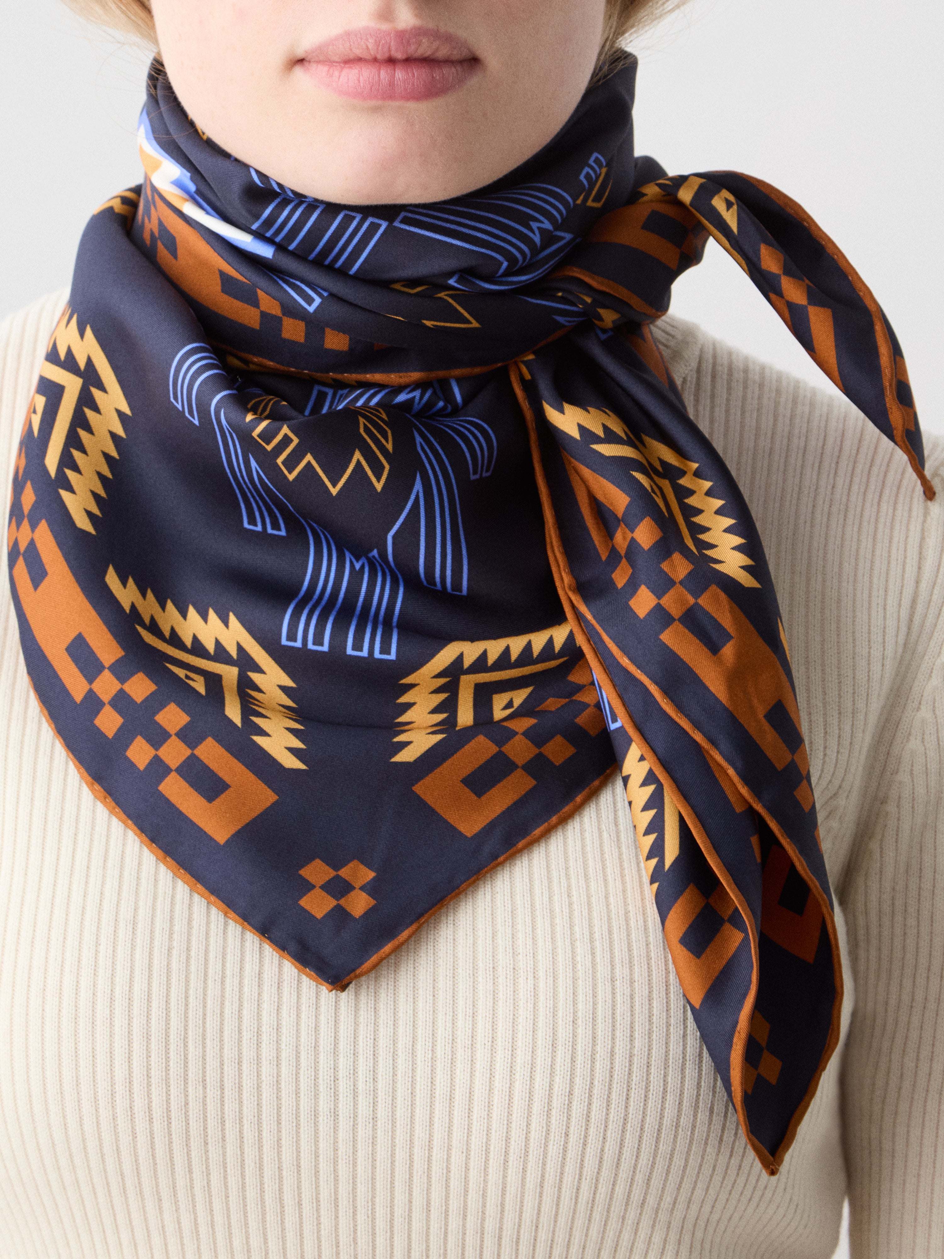 We Norwegians | FW25 | Pioneer Silk scarf_Navy Blue | Model is wearing this pure silk scarf printed with motifs inspired by Indigenous Native American art, over an off-white top, against an off-white background