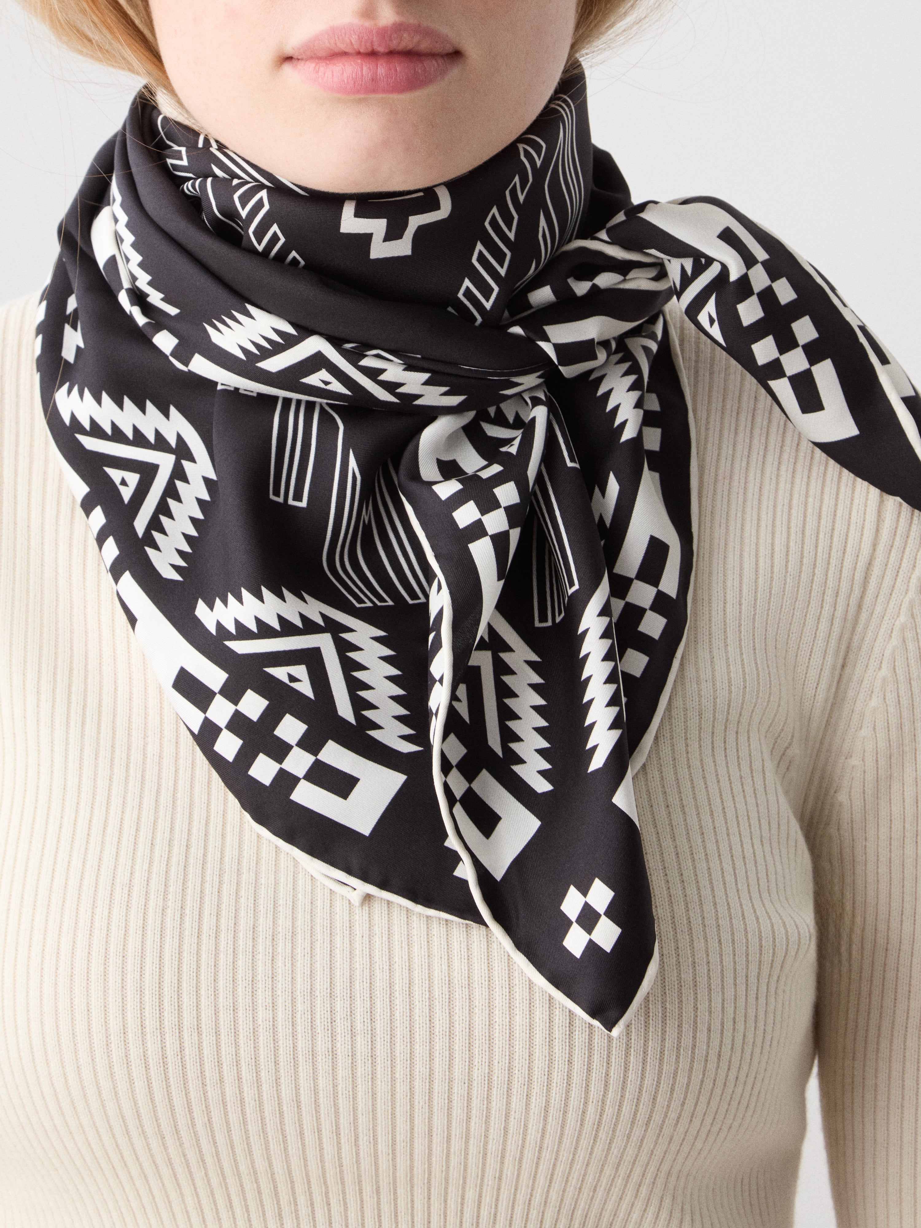 We Norwegians | FW25 | Pioneer Silk scarf_Black | Model is wearing this pure silk scarf printed with motifs inspired by Indigenous Native American art, over an off-white top, against an off-white background