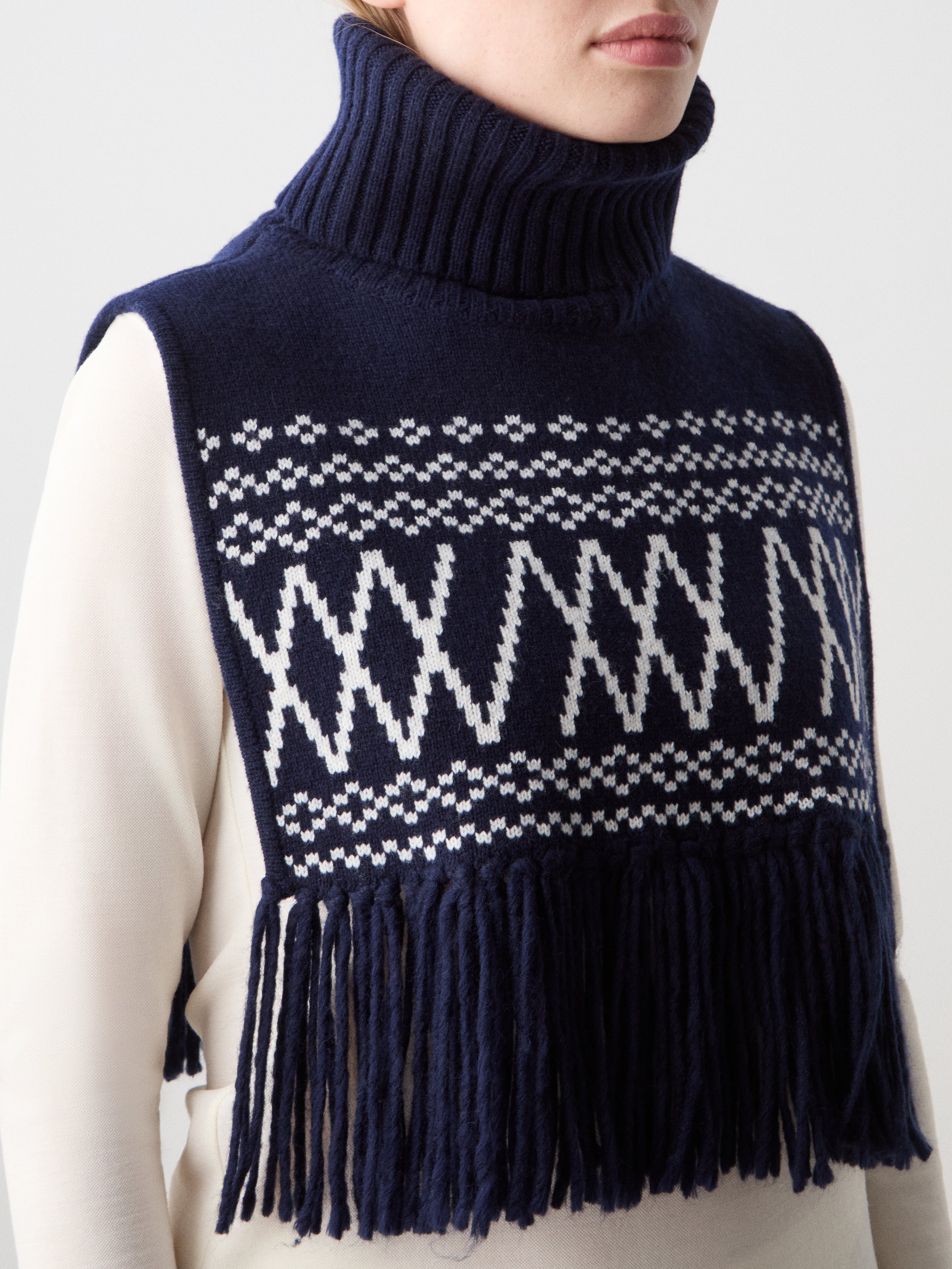 Wearing the Setesdal Bib by We Norwegians—a navy blue knit piece with white geometric patterns and fringe—over a long-sleeve white top perfectly captures classic Scandinavian style.
