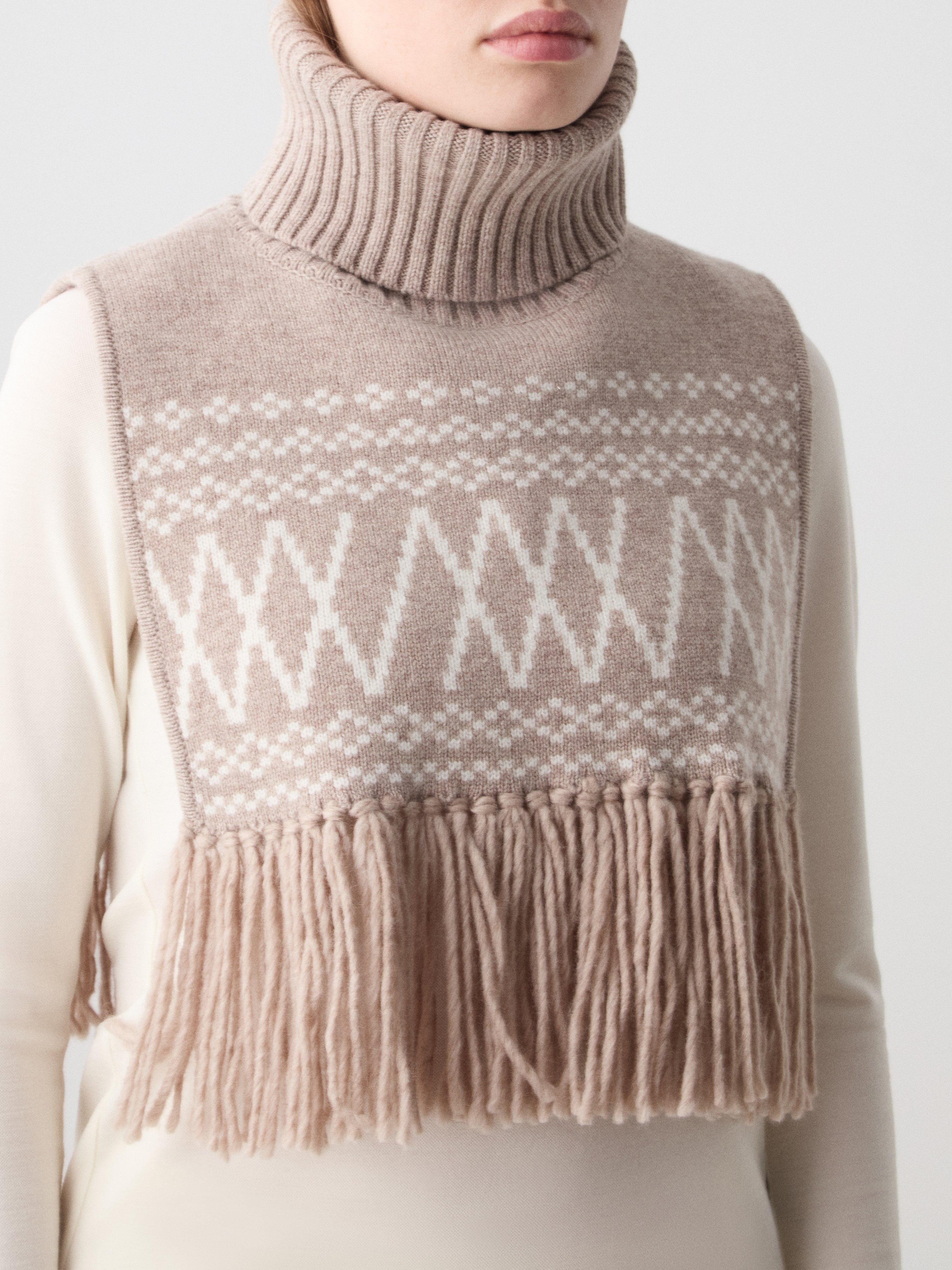 A person wears the We Norwegians Setesdal Bib—beige Merino wool with white geometric pattern and long fringe—over a cream long-sleeve top, channeling Scandinavian style with a partially visible face.