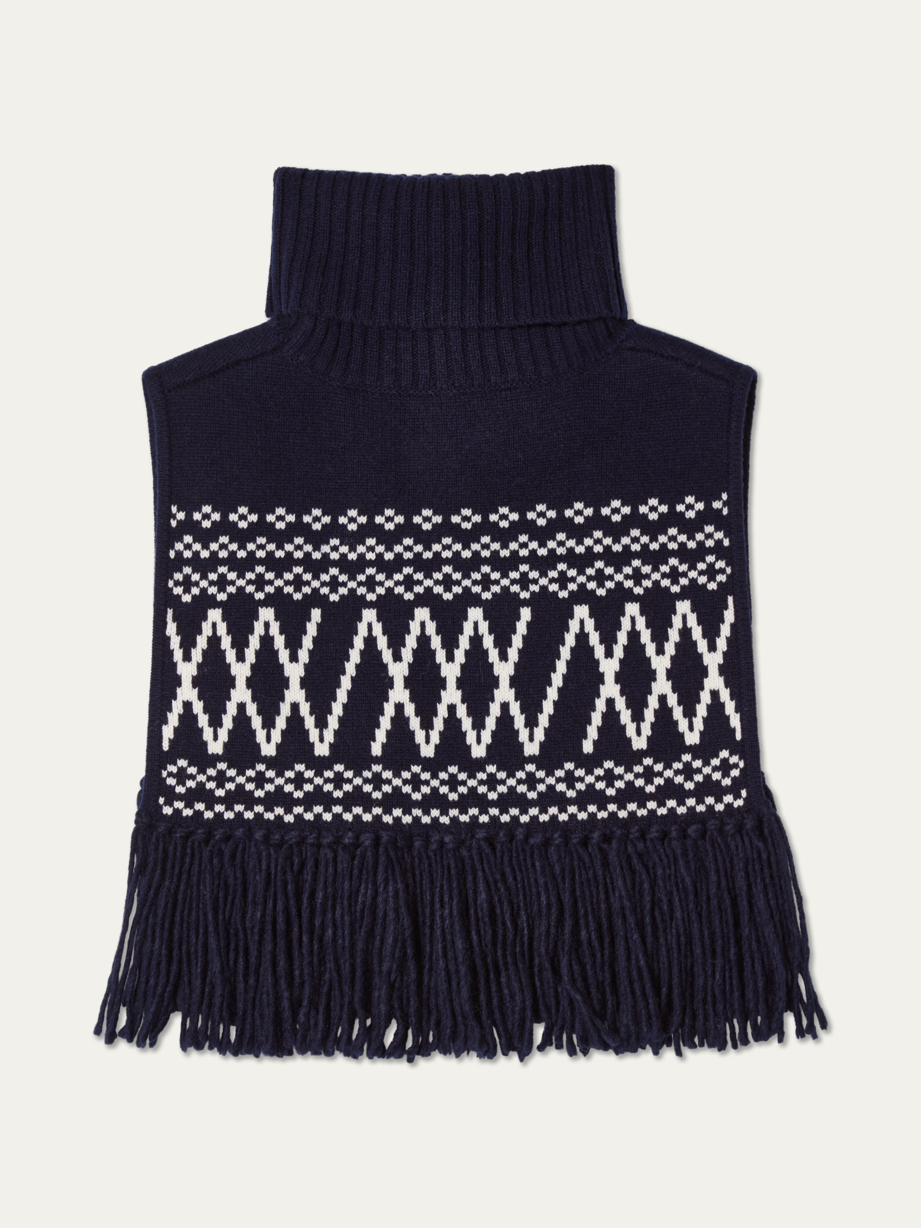 The We Norwegians Setesdal Bib is a navy blue knit dickey featuring a high ribbed turtleneck, white geometric patterns on the front, and a fringe trim, reflecting classic Scandinavian style.