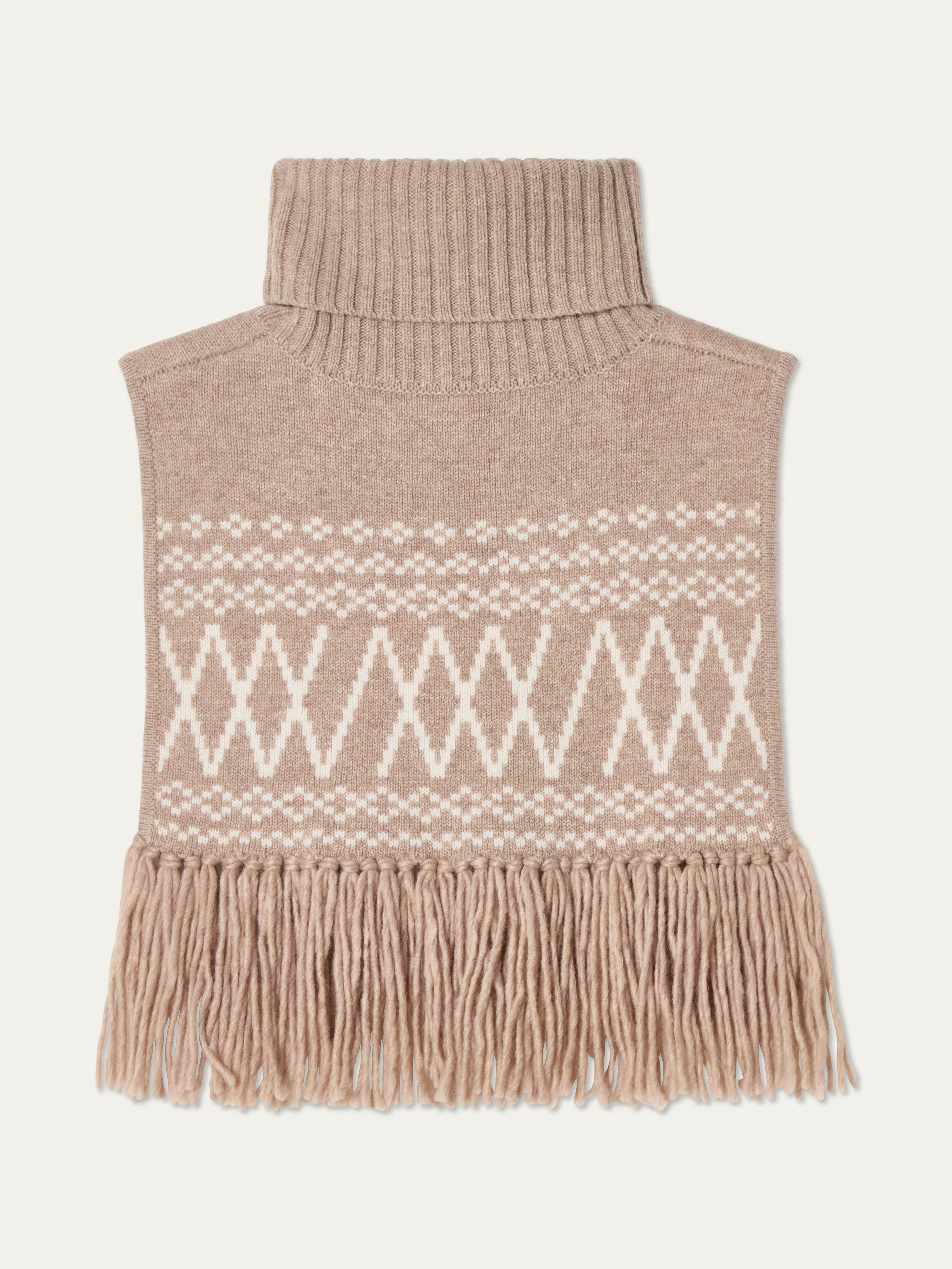 The Setesdal Bib by We Norwegians is a beige Scandinavian-style knit turtleneck dickey with a white geometric pattern and long fringes along the bottom, shown laid flat on a light background.