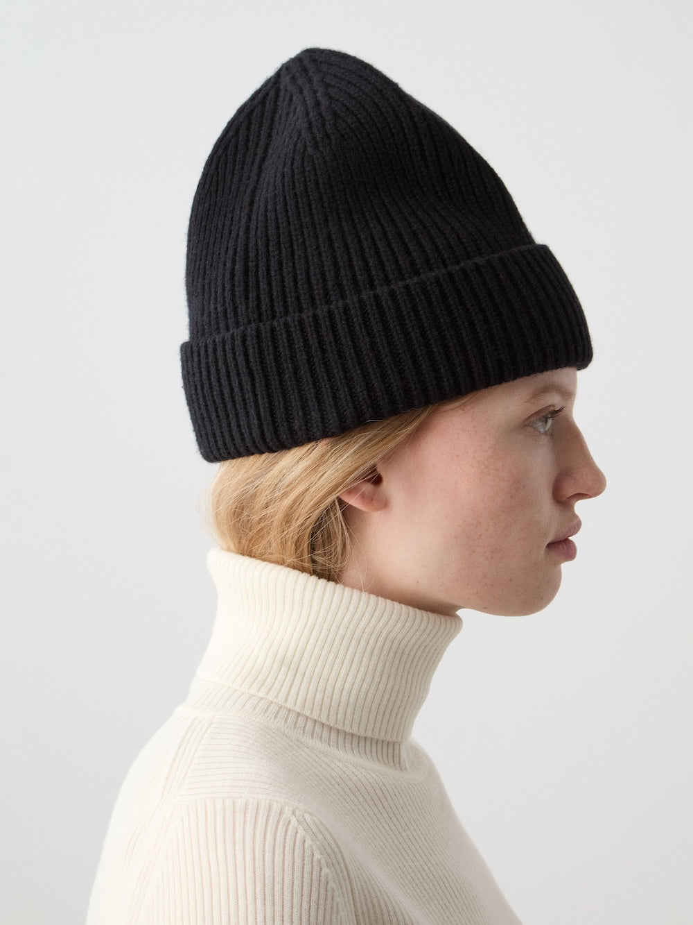 A light-skinned, blonde person wears the black We Norwegians Dal Hat and a cream ribbed turtleneck sweater, facing right against a plain light background, showcasing effortless Scandinavian style.