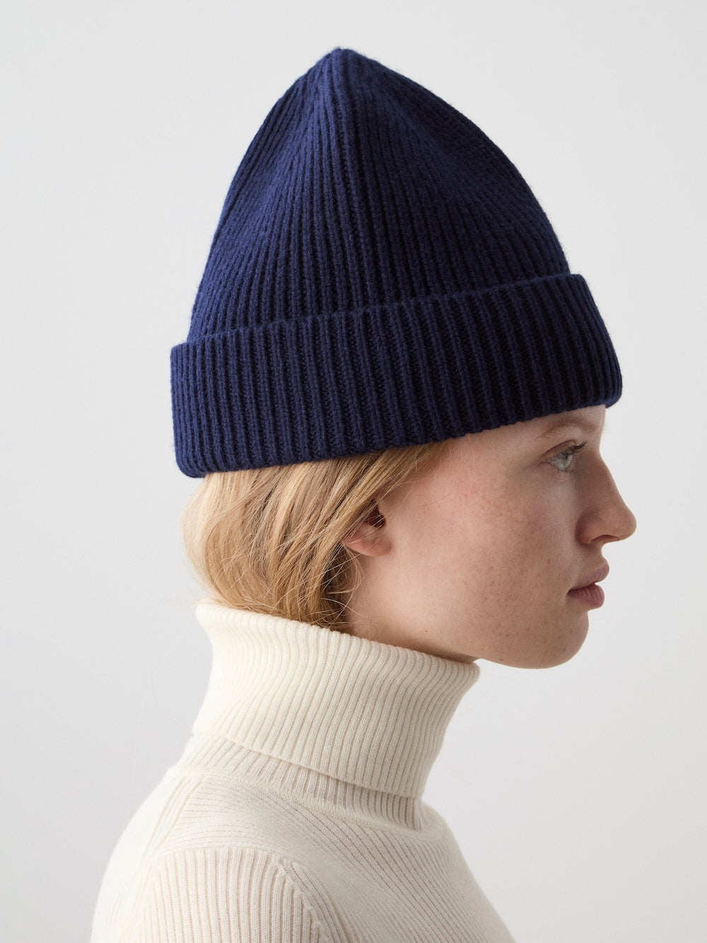 A light-skinned, blonde-haired person in profile, wearing the We Norwegians Dal Hat in navy Merino wool and a cream turtleneck sweater, stands against a plain light background, capturing understated Scandinavian style.