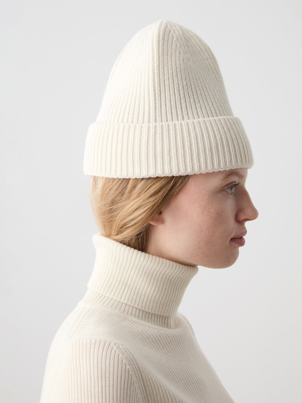 A person with light skin and blonde hair wears the We Norwegians Dal Hat—ribbed, white Merino wool—and a matching cream turtleneck sweater, capturing minimalist Scandinavian style against a plain light background. Profile view, facing right.