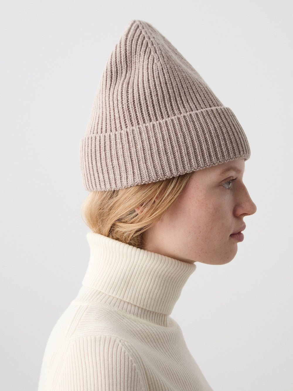 A light-skinned, blond-haired person wears the beige Dal Hat by We Norwegians, featuring a pointed top and wide cuff, paired with a cream ribbed turtleneck for a classic Scandinavian look against a simple light background.