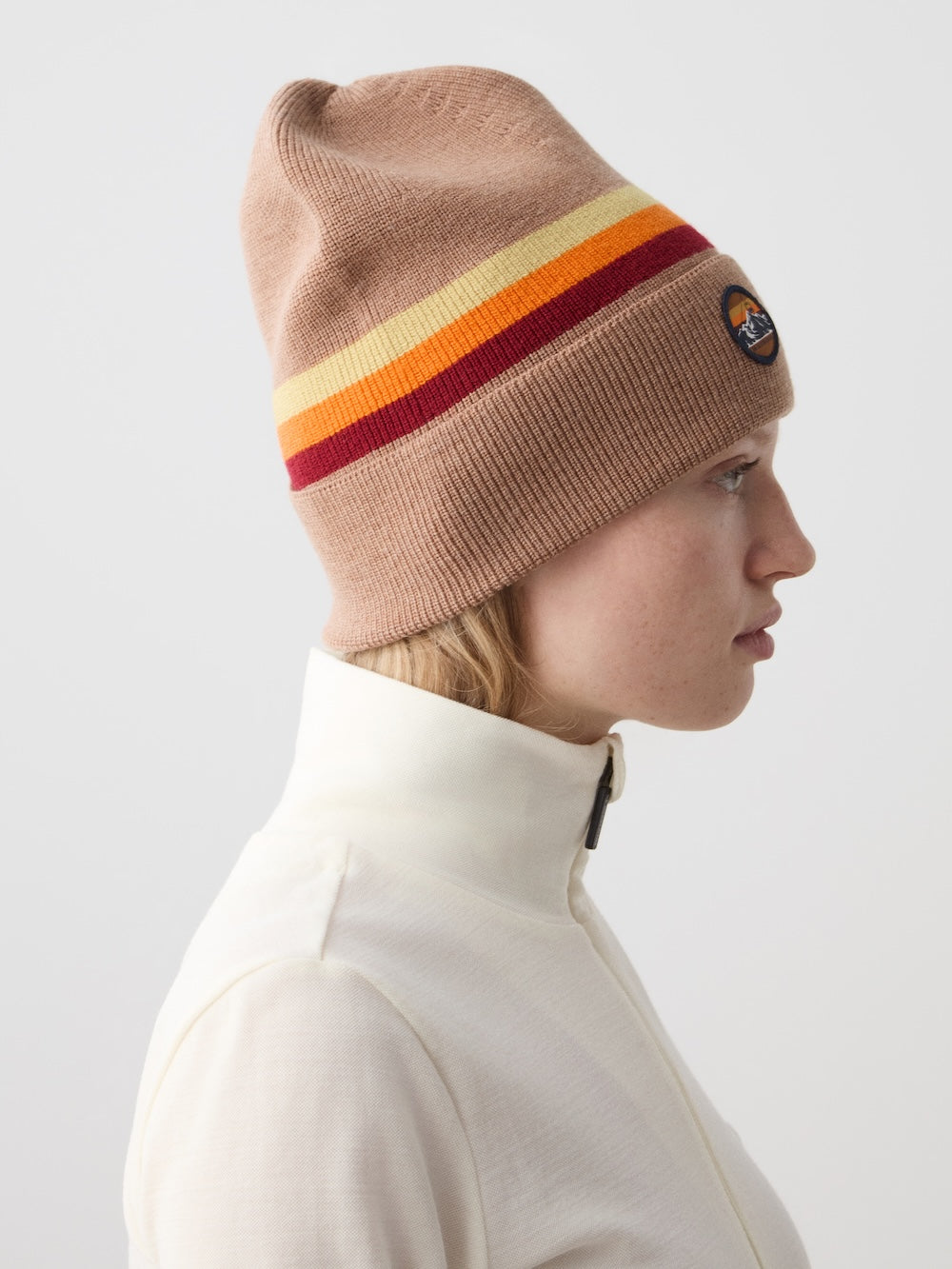 A person in profile wears the We Norwegians FreeBird Hat in beige with orange and red rainbow stripes and a circular patch, paired with a white zip-up top, against a plain light background.