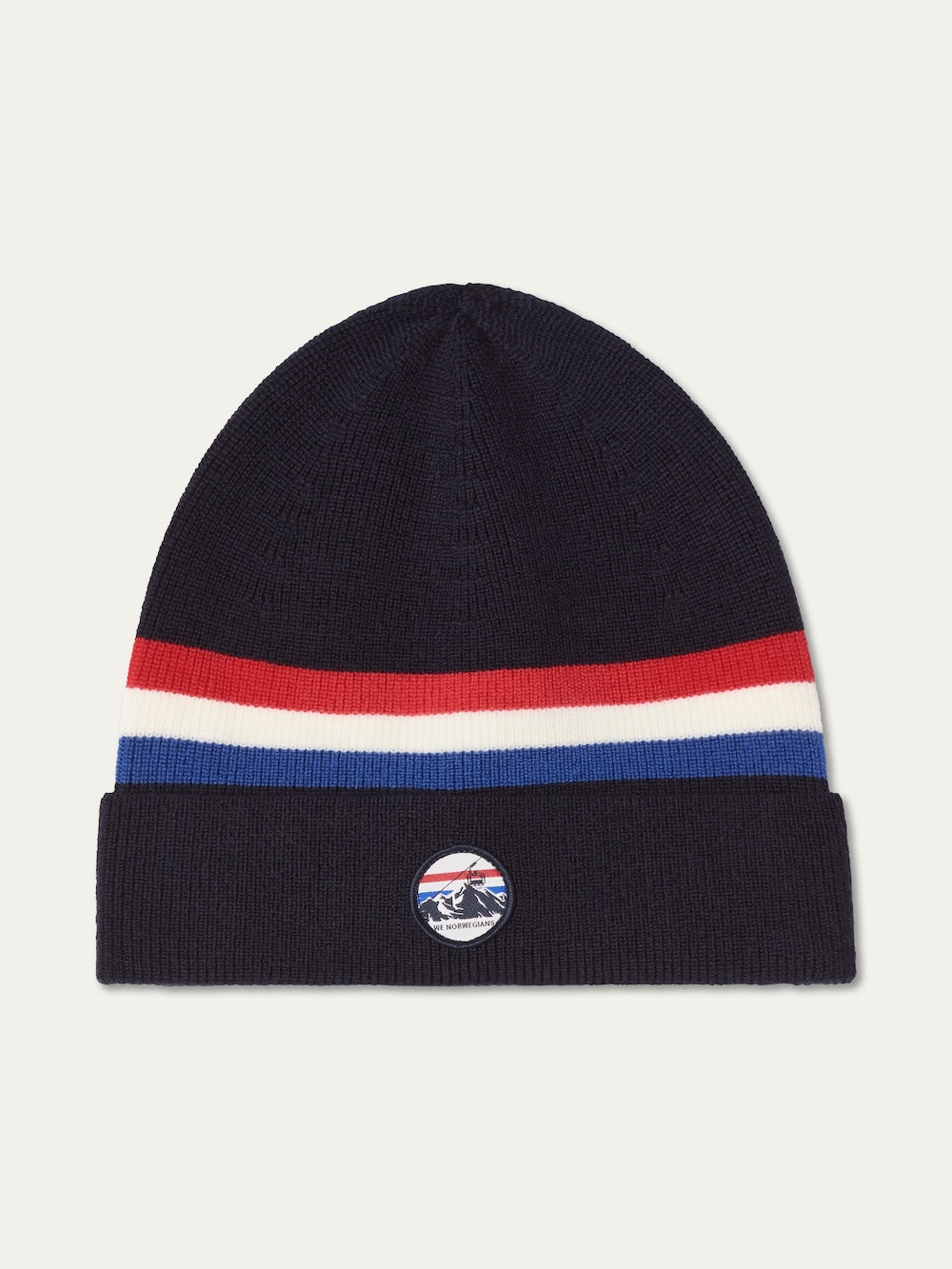 The We Norwegians FreeBird Hat is a navy blue merino wool beanie with red, white, and blue stripes, a folded brim, and a round patch featuring a mountain design.