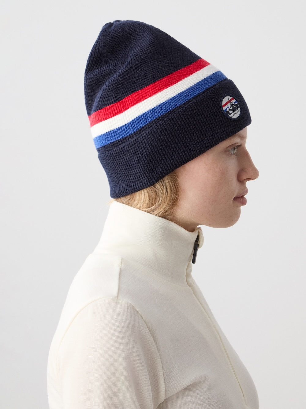 A person in a cream zip-up top is shown in profile wearing the We Norwegians FreeBird Hat, a navy beanie featuring red, white, and blue stripes with a circular side patch. The background is plain and light-colored.