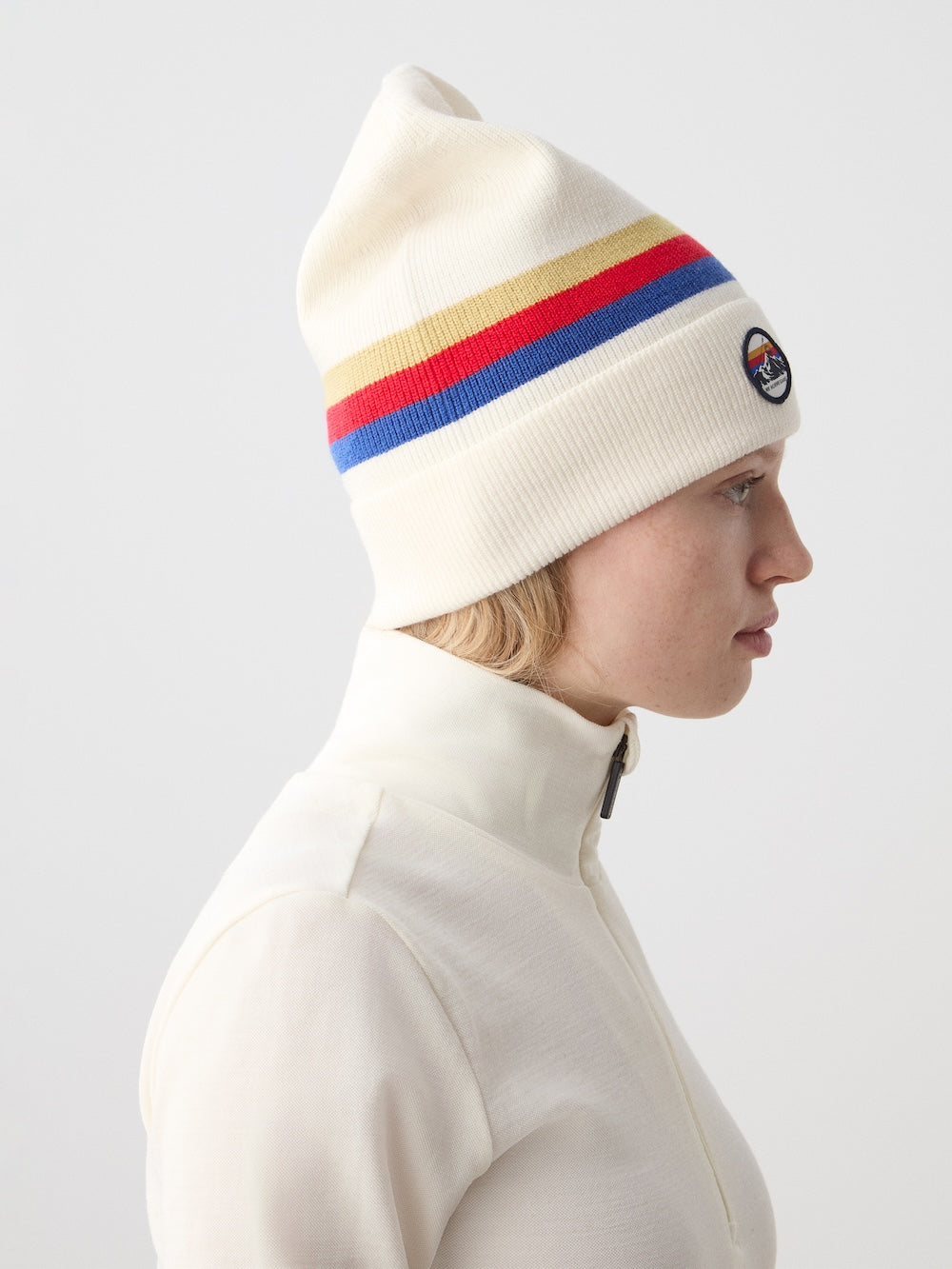 A person in a cream zip-up wears the We Norwegians FreeBird Hat—a cream merino wool hat with rainbow stripes and a round patch—seen in profile against a plain light background.